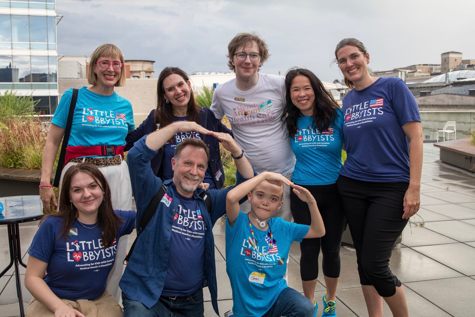 Group photo of seven Little Lobbyists parents, all wearing LL t-shirts, on the rooftop plaza of the MLK, Jr., Library, with advocate Matthew Cortland. The skyline of Washington, DC, is behind them. Photo credit: Tirrea Billings