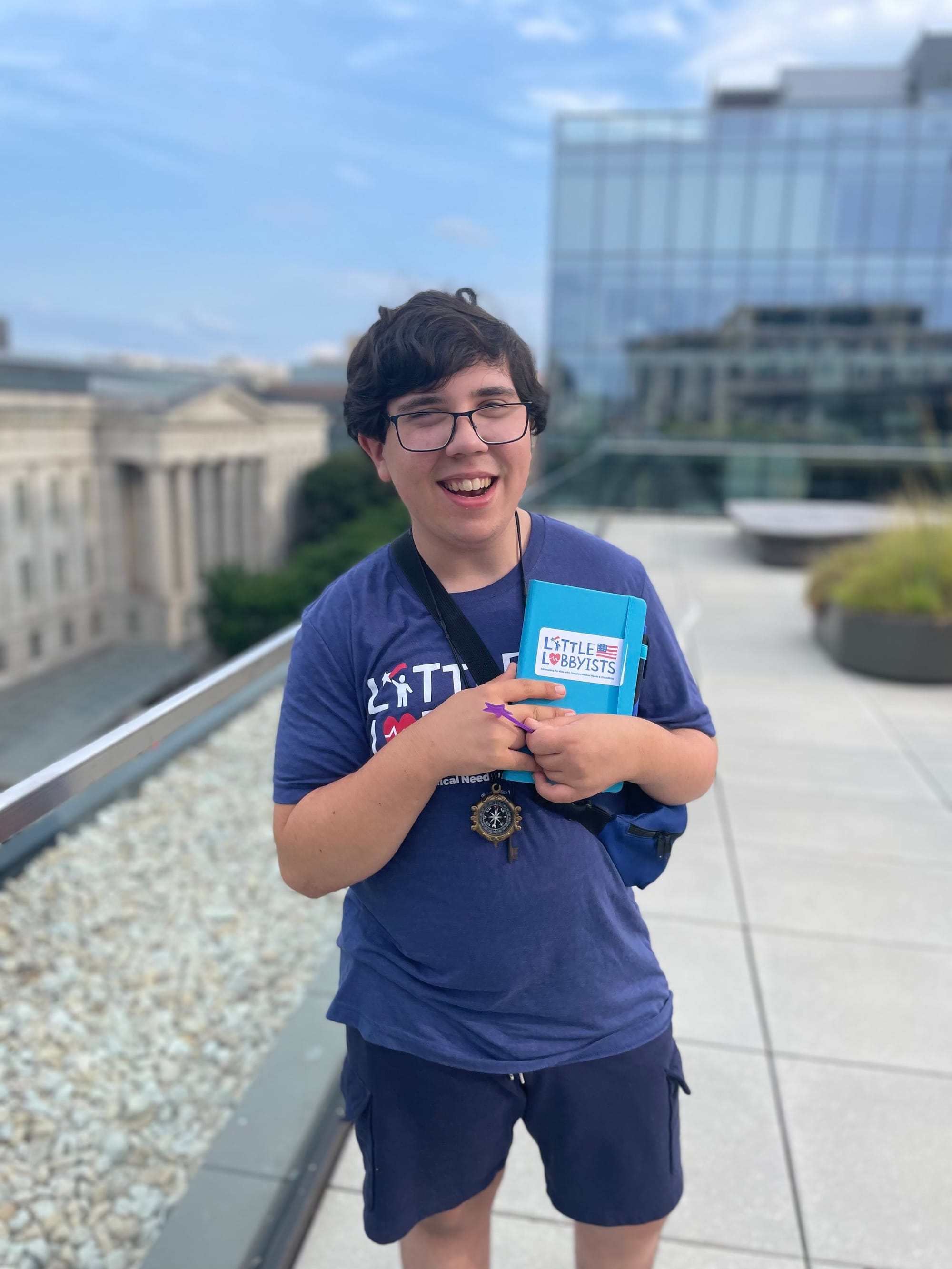 Little Lobbyist Simon, wearing a blue LL t-shirt and holding a notebook with the LL logo, poses on the rooftop plaza of the library.