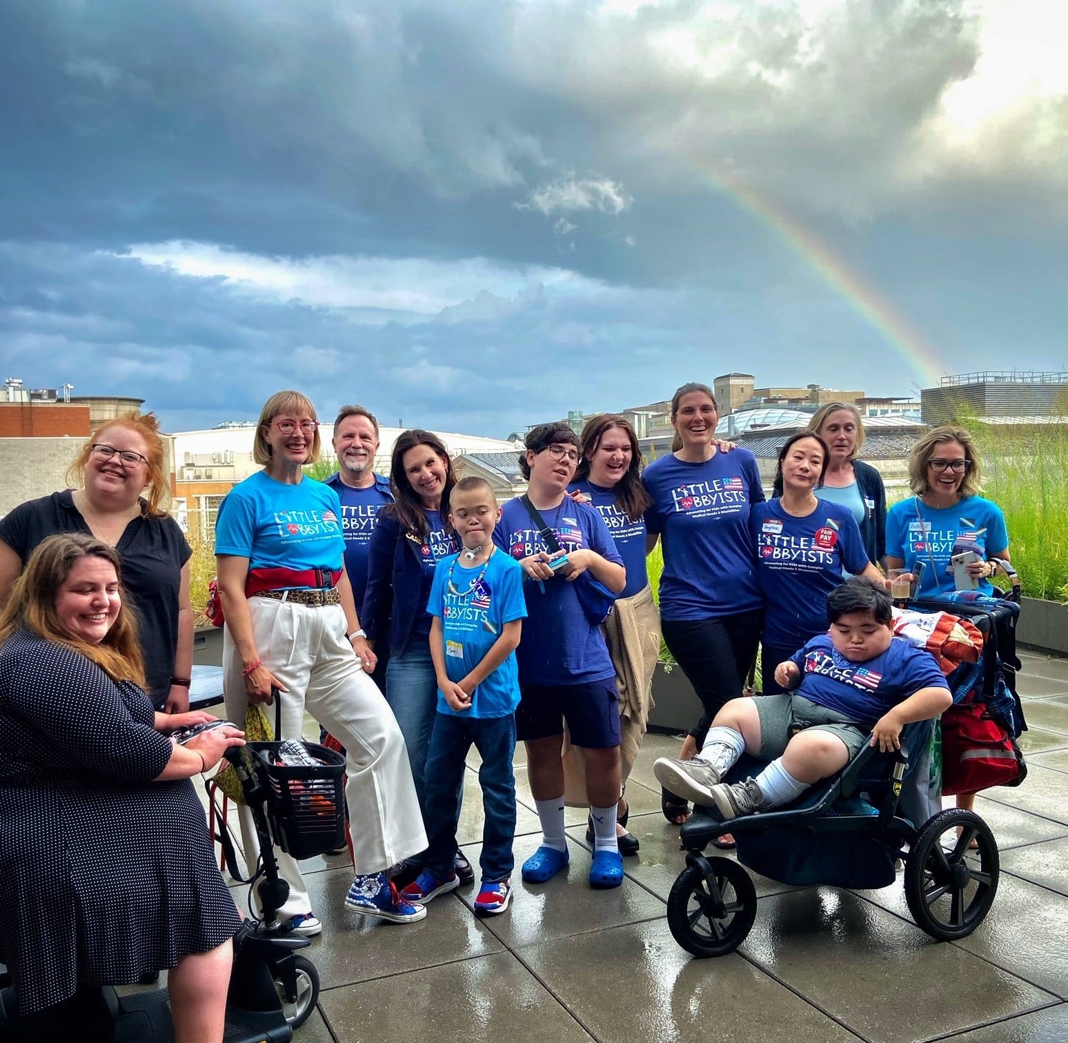 Large group photo of disability activists, Little Lobbyists parents and children, some standing, some using wheelchairs, on the rooftop plaza. Behind them is the city skyline, a bank of heavy clouds, and a rainbow.