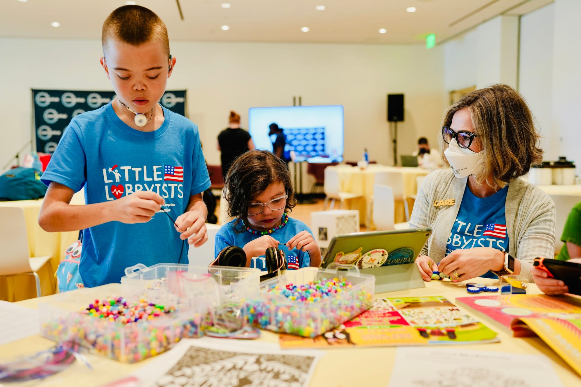 Little Lobbyists James and Louisa with LL parent Andrea. They are at the summit at a large table working on a beading project. Andrea is wearing a face mask. Photo credit: Rah Studios
