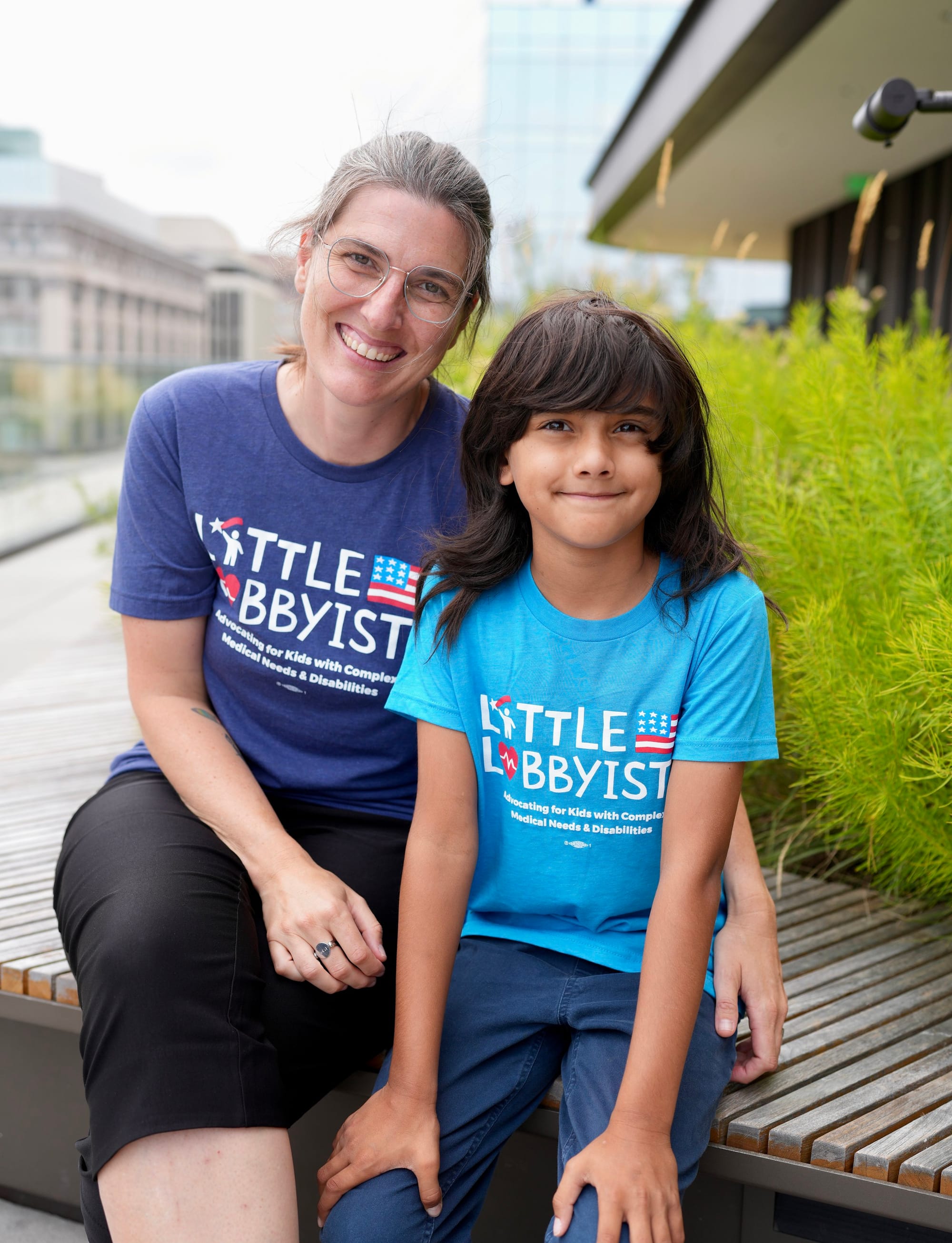 Little Lobbyists Ali and Ethan wear dark blue and turquoise LL t-shirts, respectively. They pose on the rooftop plaza of the library. Visible behind them is the city skyline, decorative foliage in a planter, and a roof overhang. Photo credit: Rah St