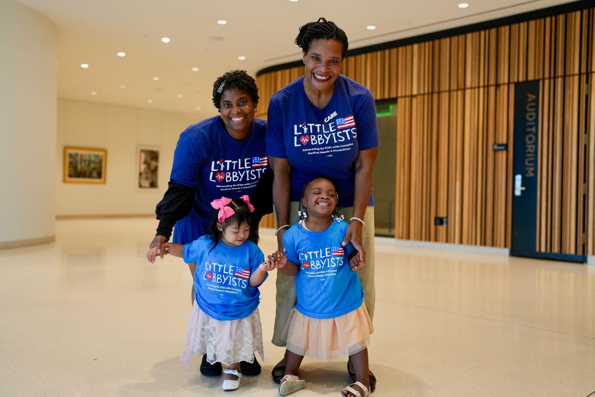 Little Lobbyists Danielle and Destiny pose with friends. Both moms and their toddler daughters wear LL t-shirts. They are inside the conference area at the library. Photo credit: Rah Studios
