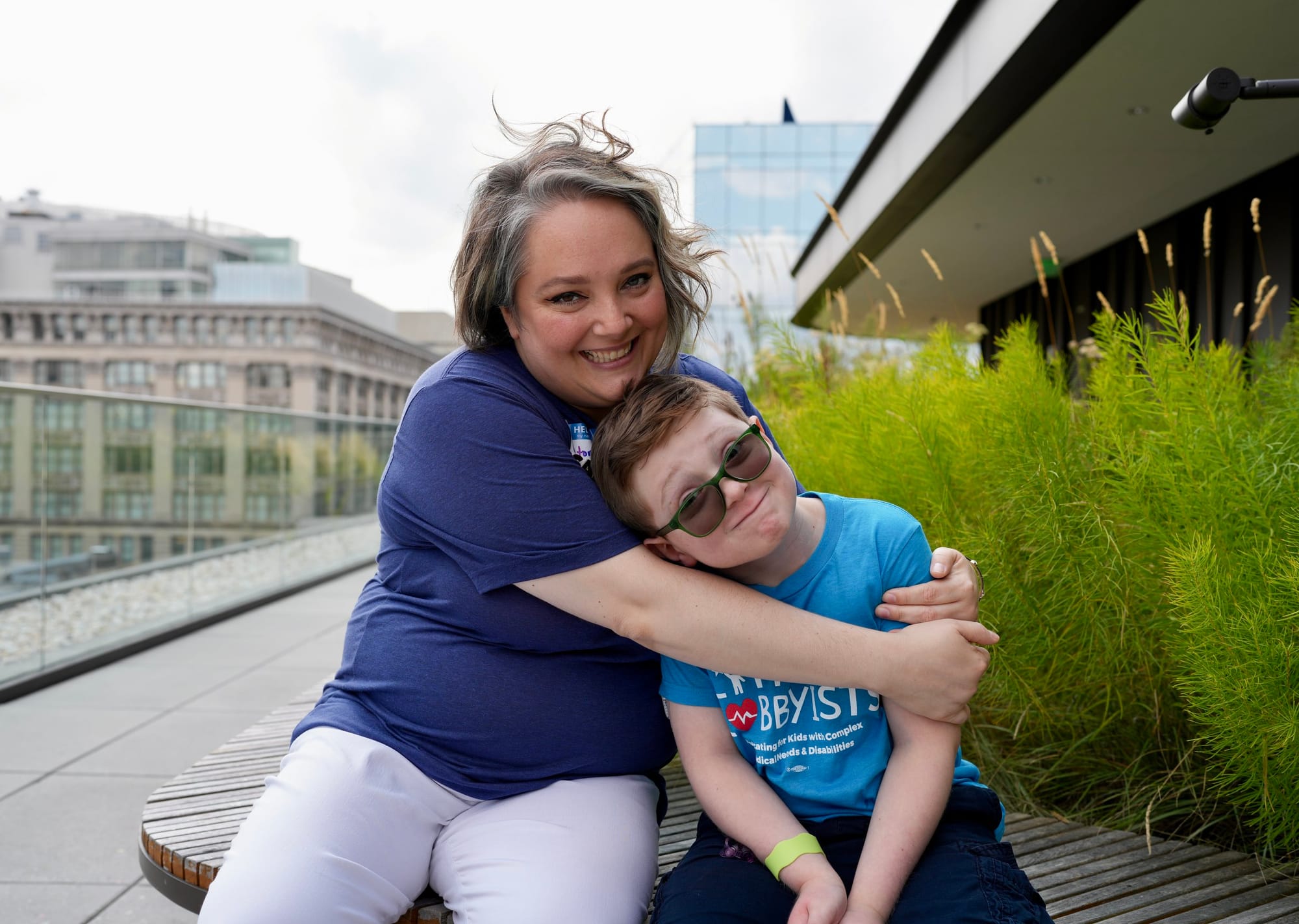 Little Lobbyists Victoria and Cole sit outside on the library rooftop plaza. Victoria has her arms around Cole, who leans into her. They wear LL t-shirts and the city skyline is behind them. Photo credit: Rah Studios