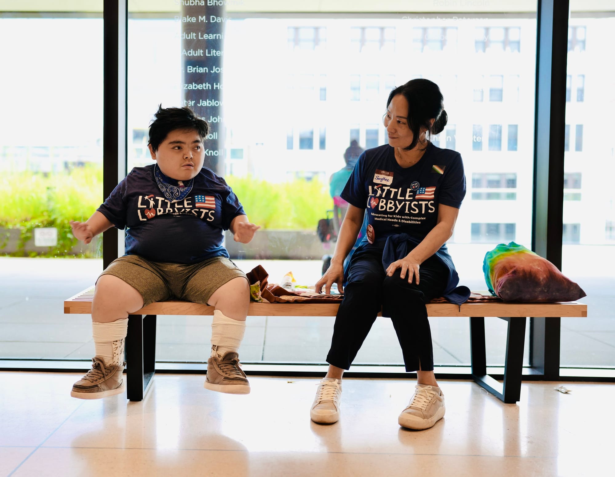 Little Lobbyists Teddy and Sanghee sit on a long bench inside the library, with plate glass windows behind them showing the plaza and buildings. They are wearing LL t-shirts. Photo credit: Rah Studios
