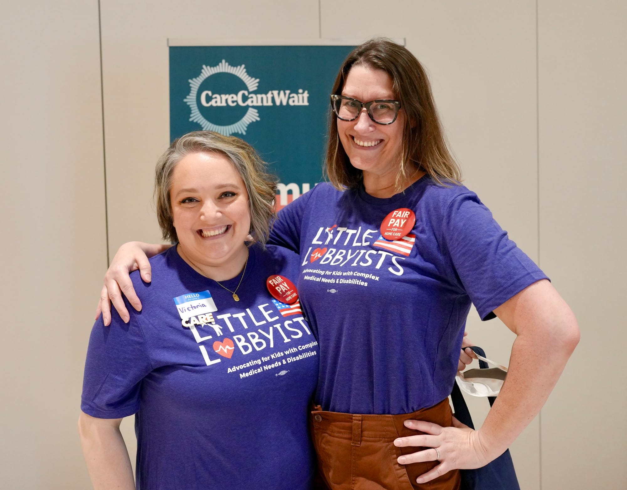 Little Lobbyists Victoria and Meghann pose with their arms around each other. They wear LL t-shirts and a Care Can't Wait banner is behind them. Photo credit: Rah Studios