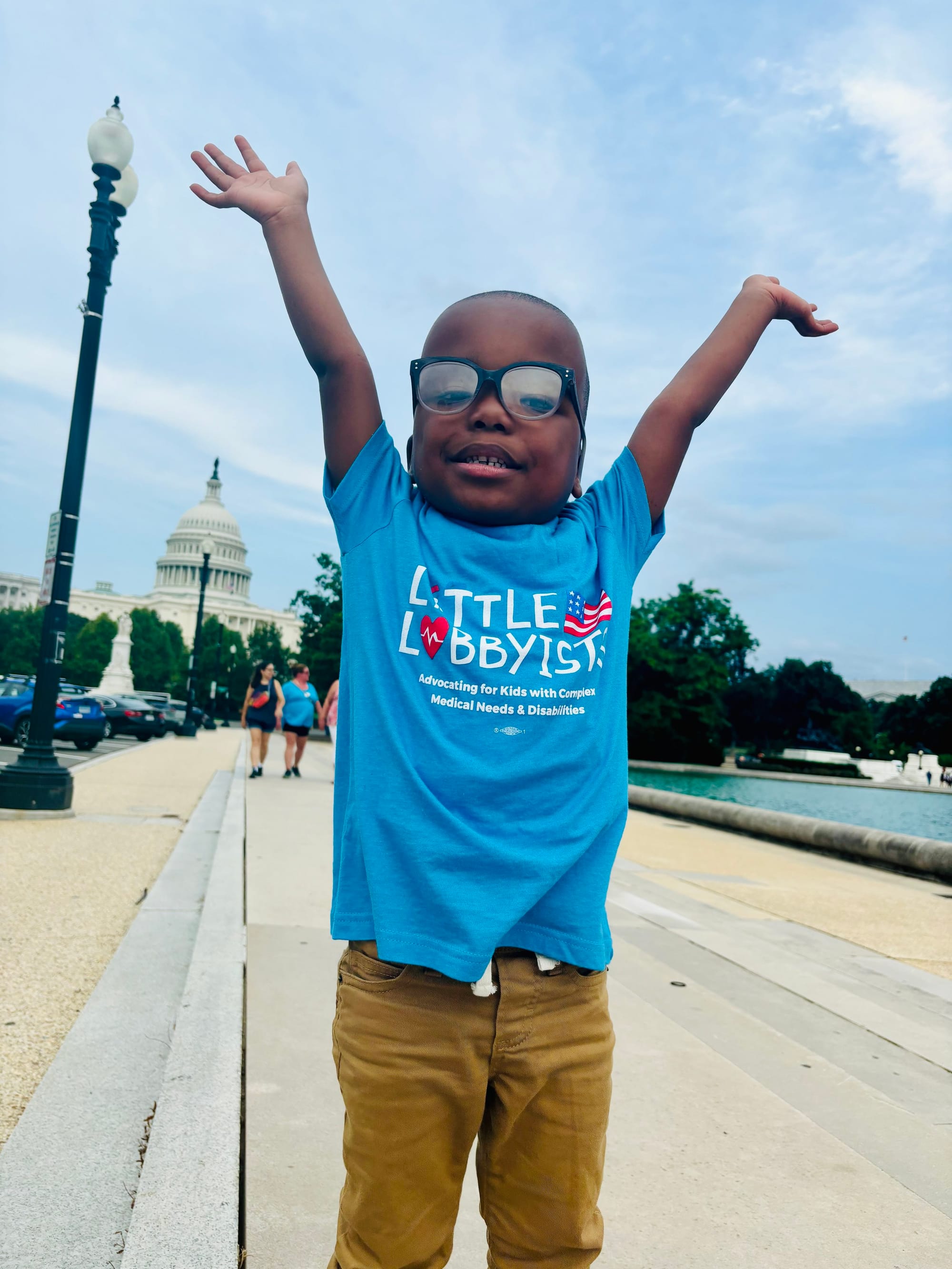 Little Lobbyist Adonise poses on a DC city street with both arms raised high. Behind him, in the distance, is the U.S. Capitol building. Adonise wears a turquoise LL t-shirt and brown pants.
