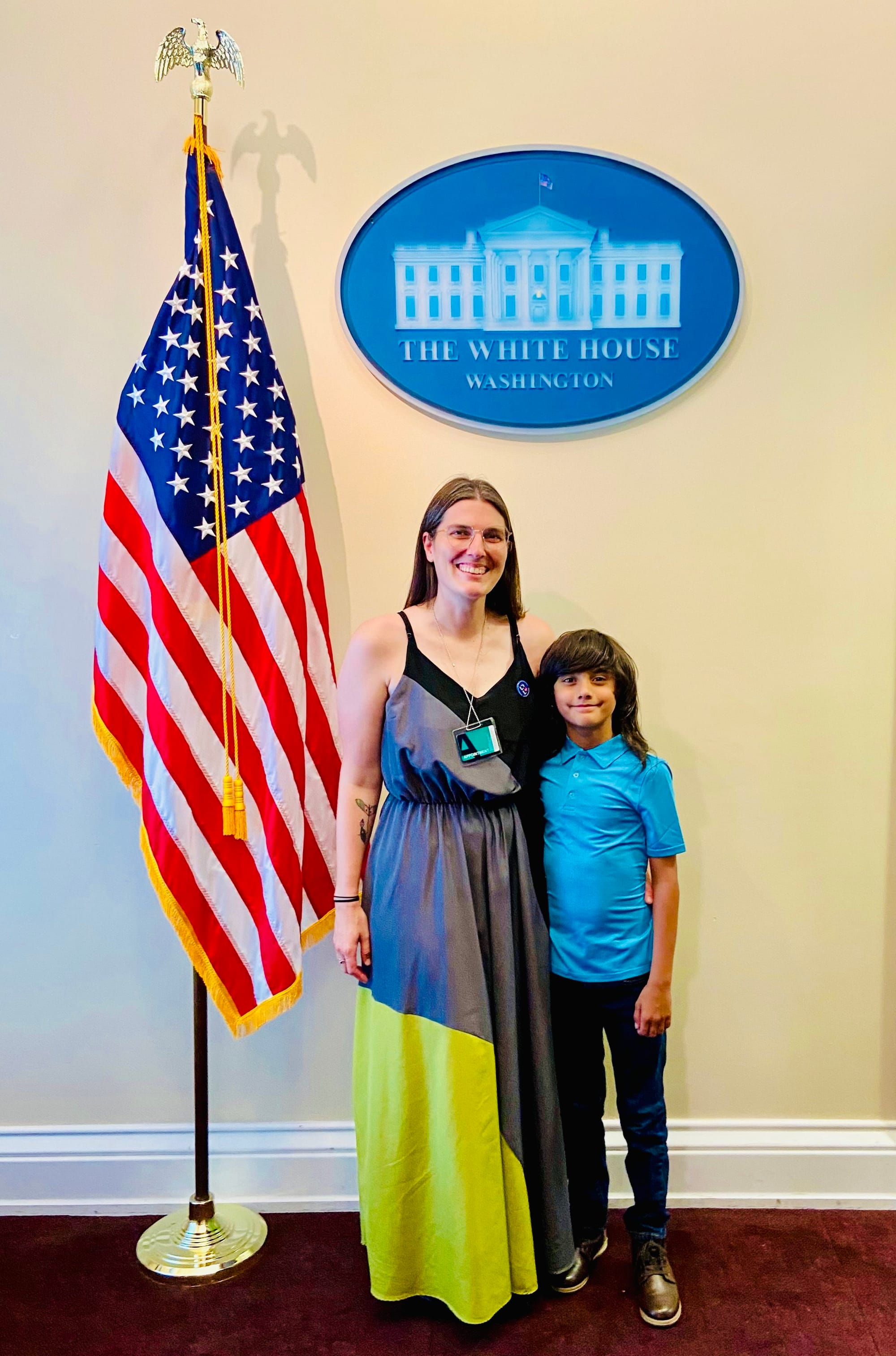 Little Lobbyists Ali and Ethan pose in the Old Executive Office building in front of a wall with the White House logo (blue & white oval) above them and a U.S. flag to the left. Ali wears a long skirt and Ethan is dressed more casually.