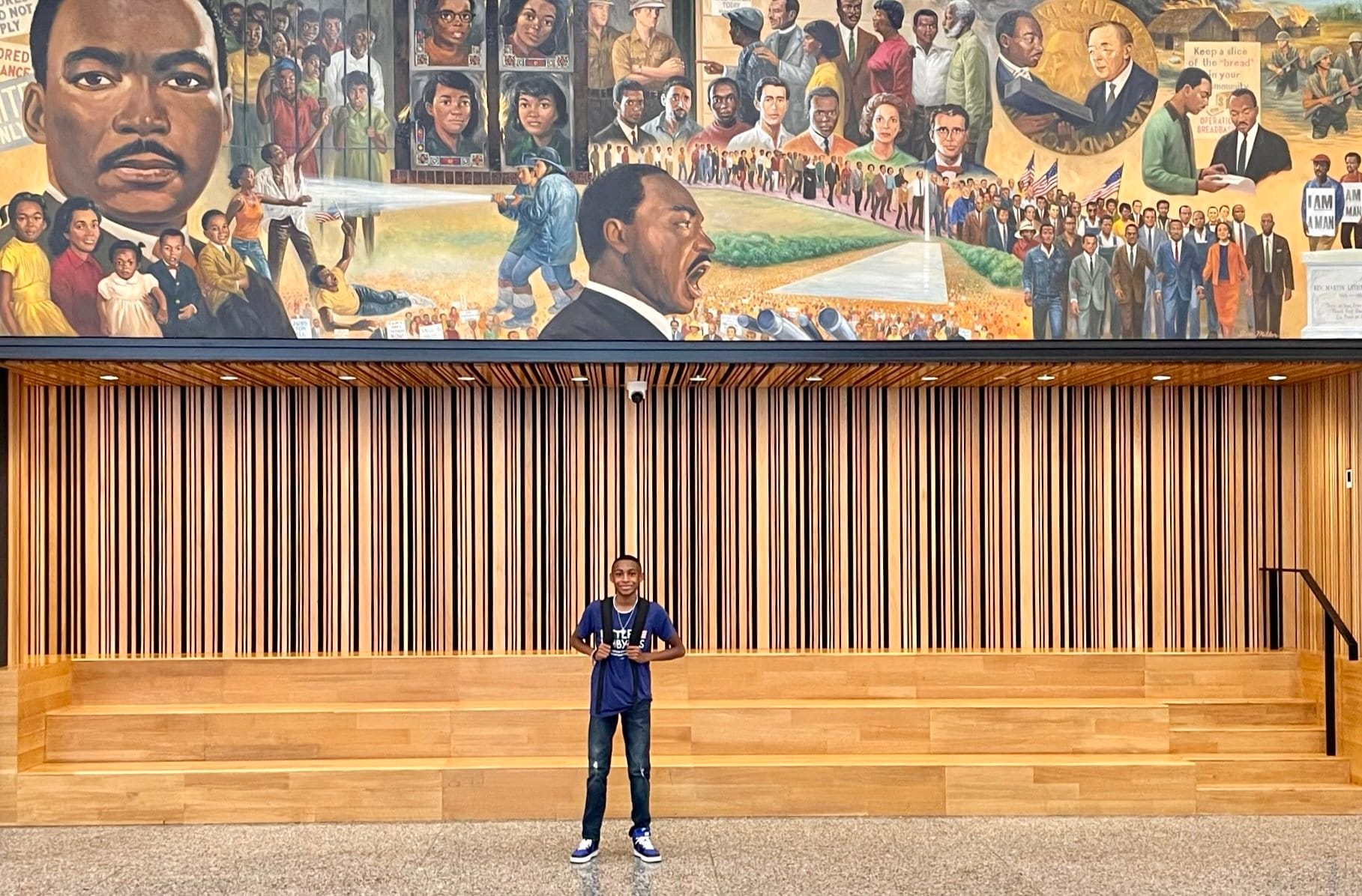 Isra'El poses--a small figure--under the giant mural at the MLK Jr Library. The mural depicts colorful scenes from the life of Martin Luther King, Jr.