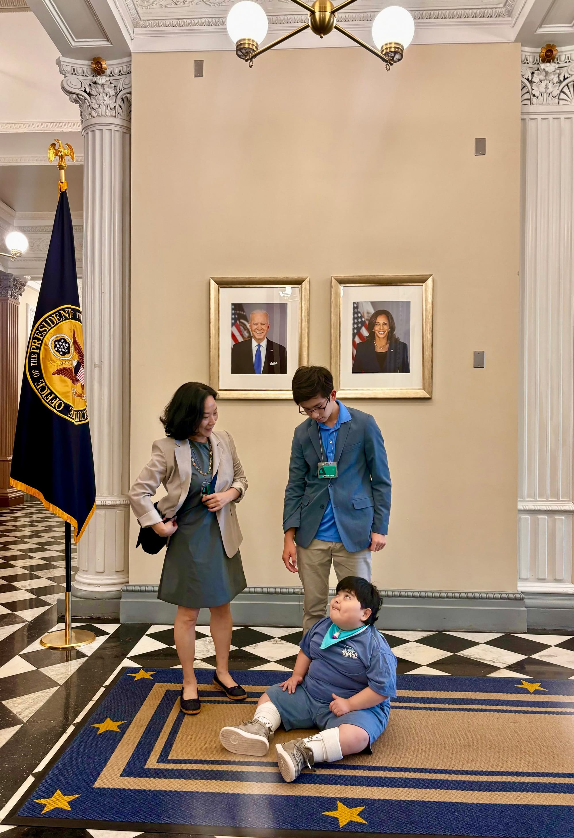 Sanghee poses with her sons Teddy and Leo in the OEB. A decorative flag is to the right. Teddy sits on a rug that partially covers a black and white tiled floor. He looks up at a decorative light fixture. Framed photos of President Biden and Vice Pr