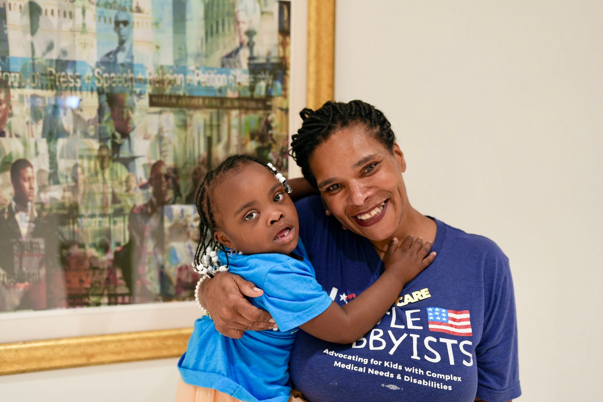 Little Lobbyists Danielle and Destiny pose in front of a framed abstract painting inside the library's conference center. They are wearing LL t-shirts and smiling. Danielle is hold Destiny, who has her arms around her mom's neck. Photo credit: Rah S