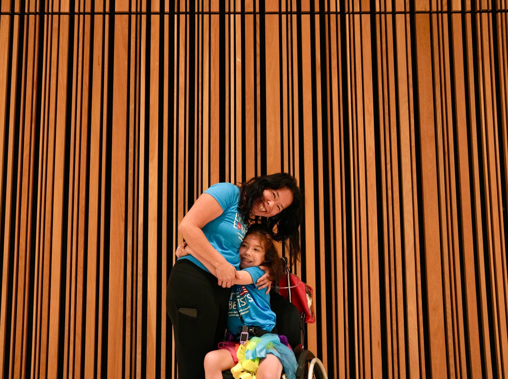 Elena and Xiomara pose in front of an elaborately wood-paneled wall (different shades of narrow wood paneling in long vertical stripes) at the MLK Jr. Library. They wear turquoise LL shirts. Elena bends down to hug Xiomara, who is seated in her whee
