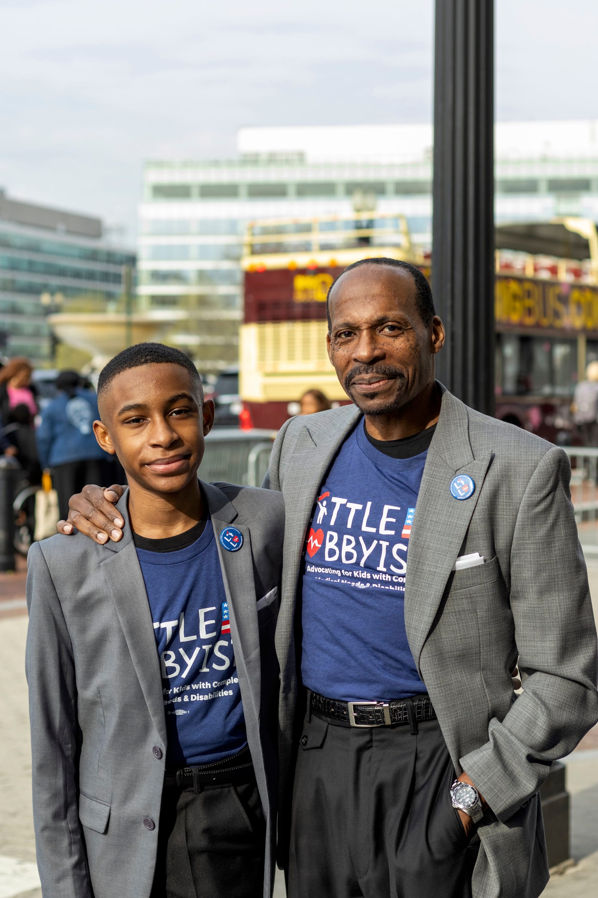 Isra'El & Rico pose outside Union Station. They wear gray suit jackets with LL pins on the lapel and LL t-shirts underneath. Both have close-cropped, tightly curled dark hair. Rico has a mustache.