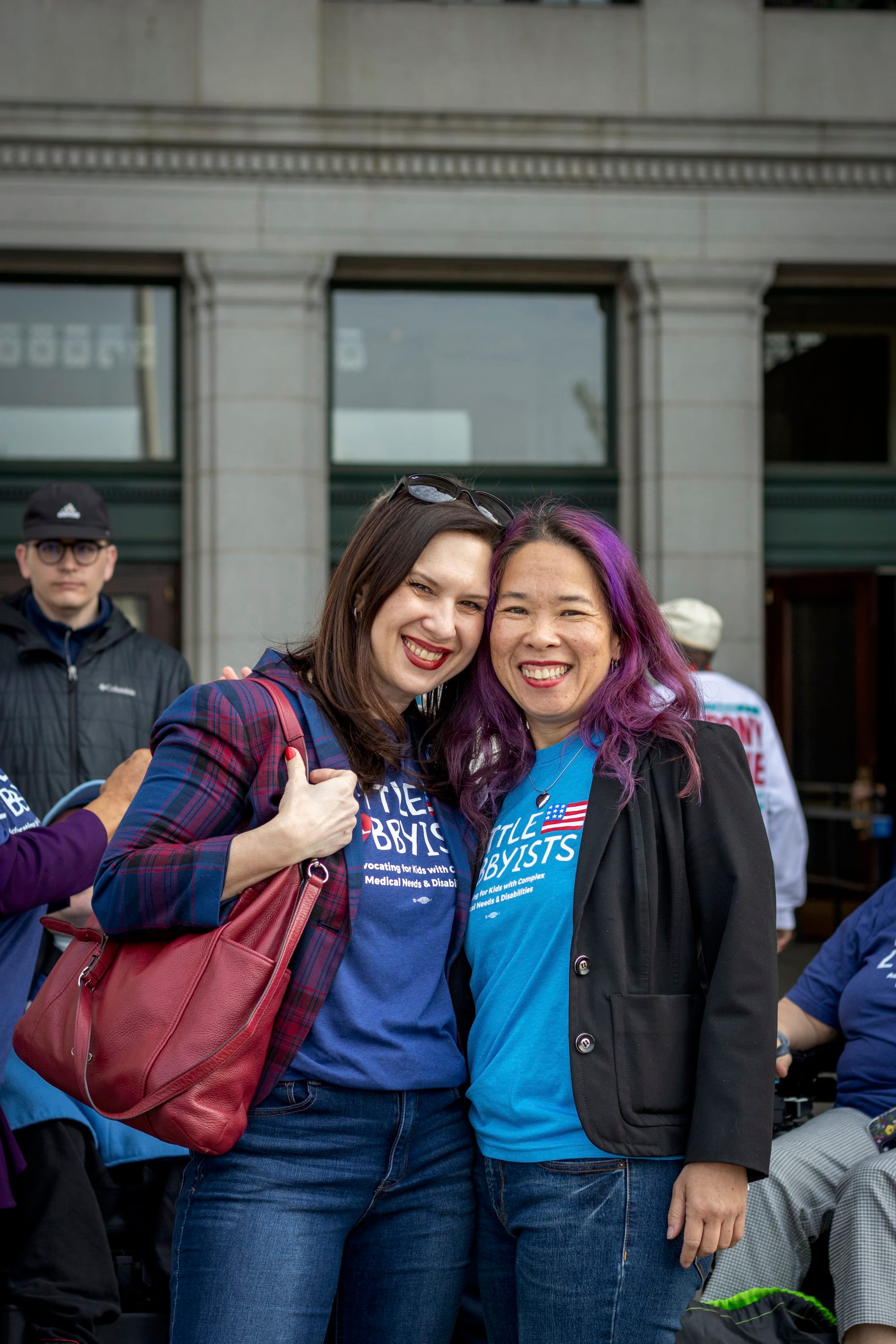 Laura and Elena pose outside the station. Laura has a large red bag over her shoulder. Elena has bright magenta streaks in her dark hair.