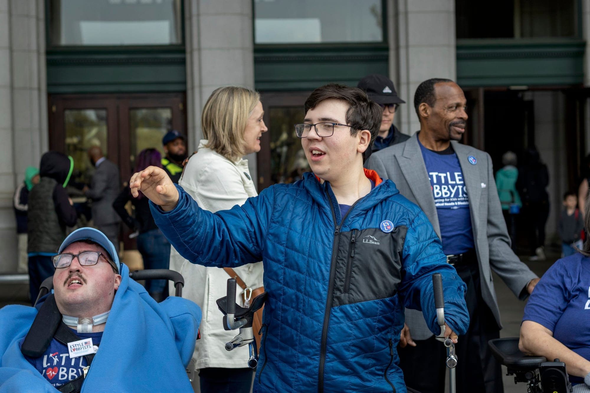 Simon stands using his walker and points to something in the distance. Rob, who sits in his wheelchair, is to the left.