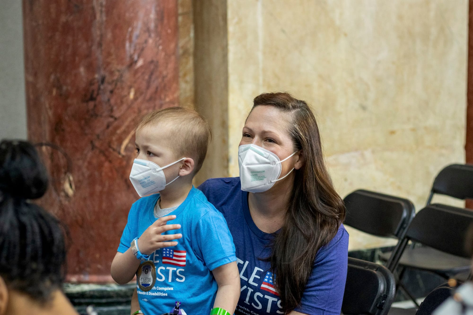 Megan holds her son Gabe in her lap inside the rally. Both are wearing N95 masks and they are seated alongside a beautiful marble wall in earth tones.