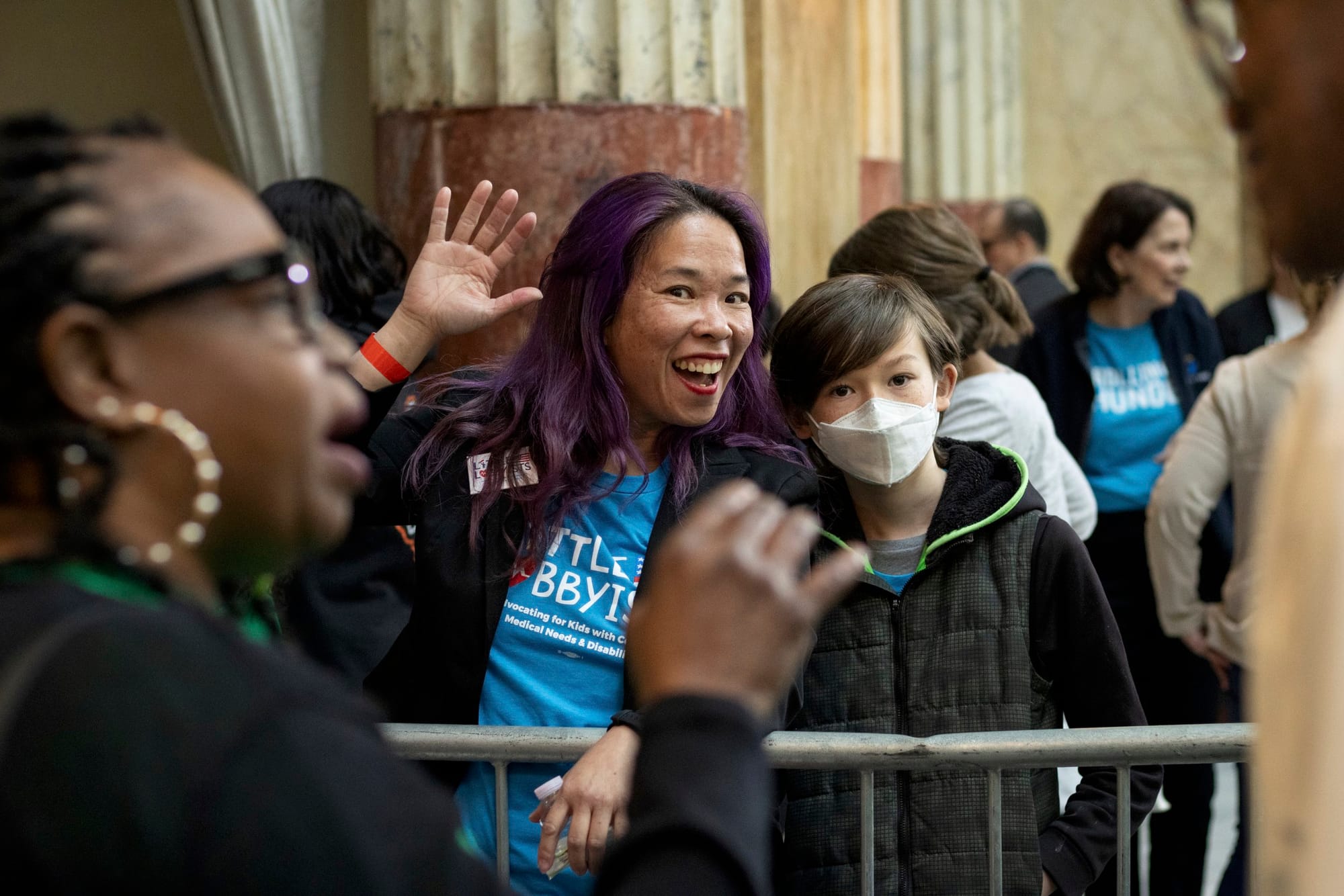 Elena and her son Khalil wave from behind a metal barrier just outside the ADA section. Khalil is wearing an N95 mask.