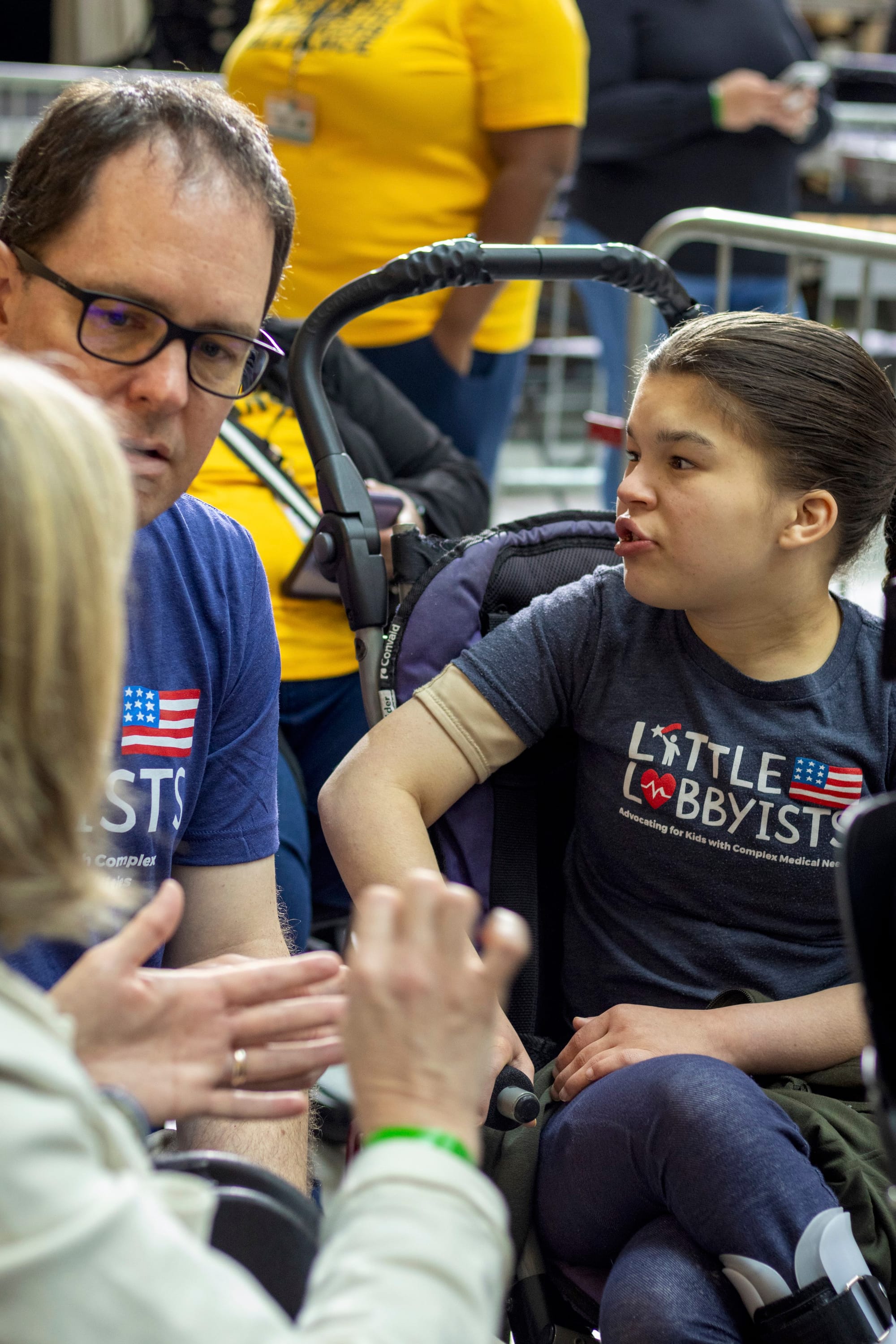 Dan and his daughter Claire are seated at the rally, talking to Jeneva, who wears an off-white coat and has her back to the camera.