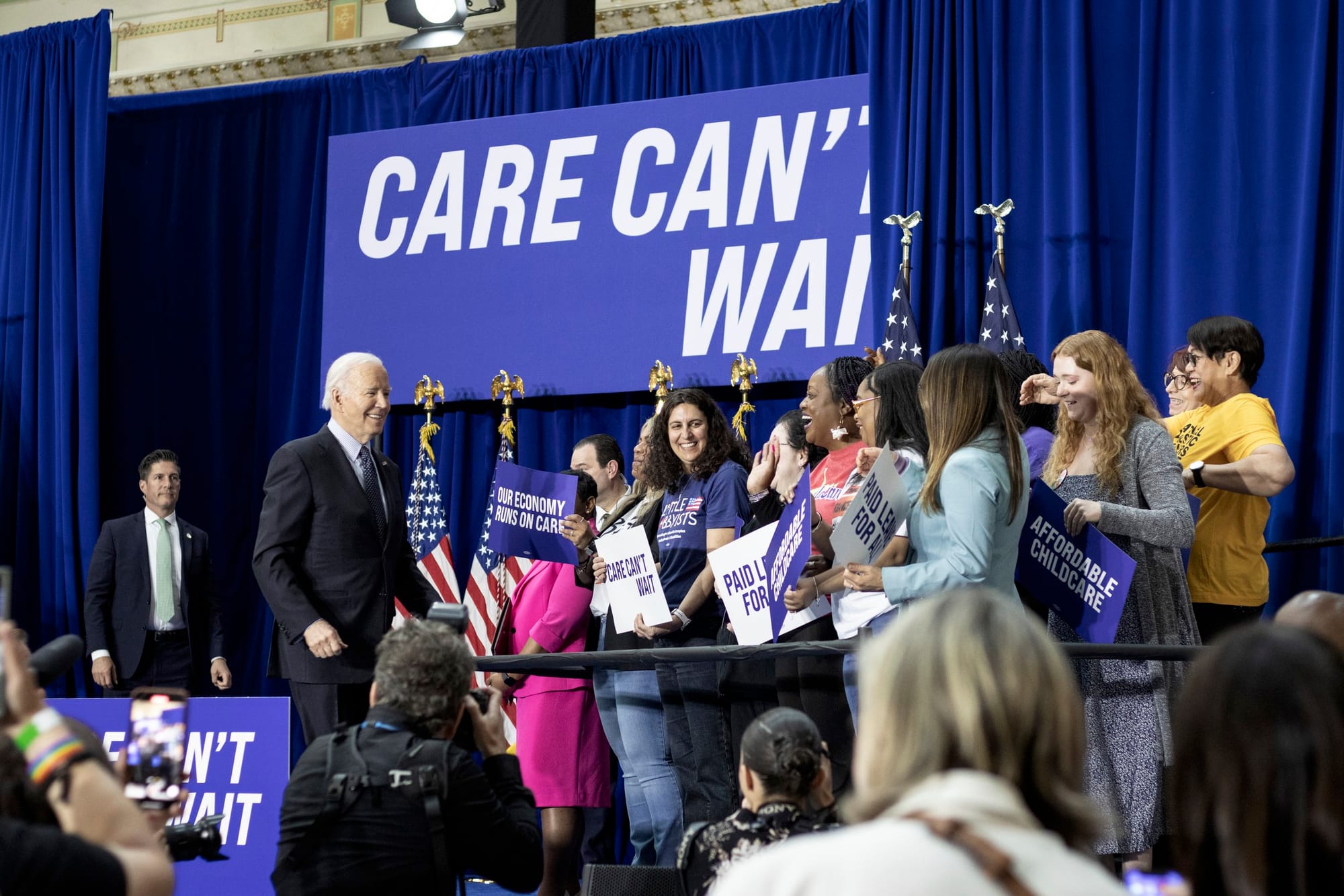 President Biden walks toward a group of caregivers to the right. They hold signs expressing support for care. Among them is Jamie, who has long, dark curly hair. Behind them is a large sign on the stage, royal blue with white lettering: Care Can’t W