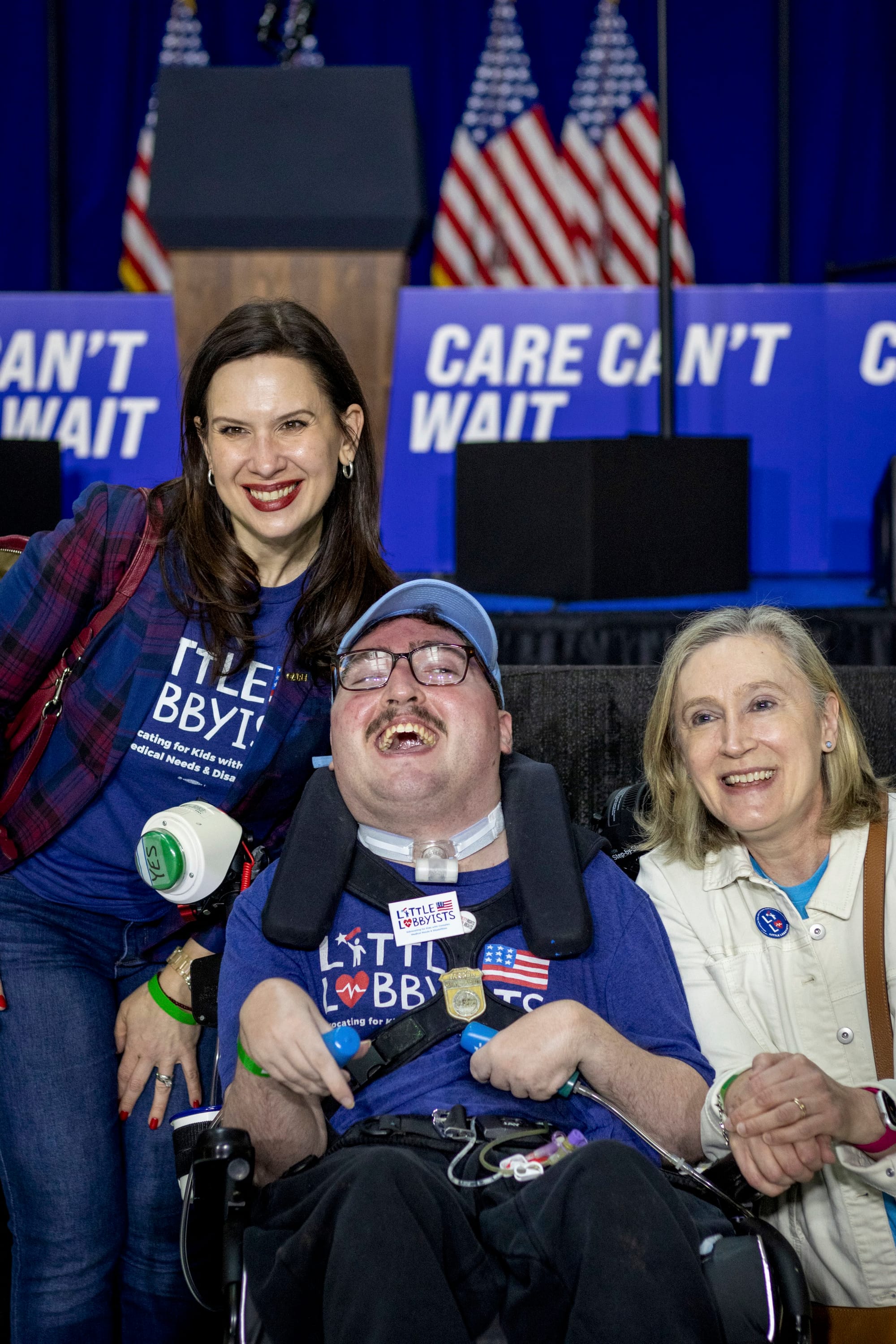 Laura and Jeneva pose on either side of Rob, who is smiling broadly from his wheelchair and wearing his LL baseball hat and his Clark-Kent-style glasses.
