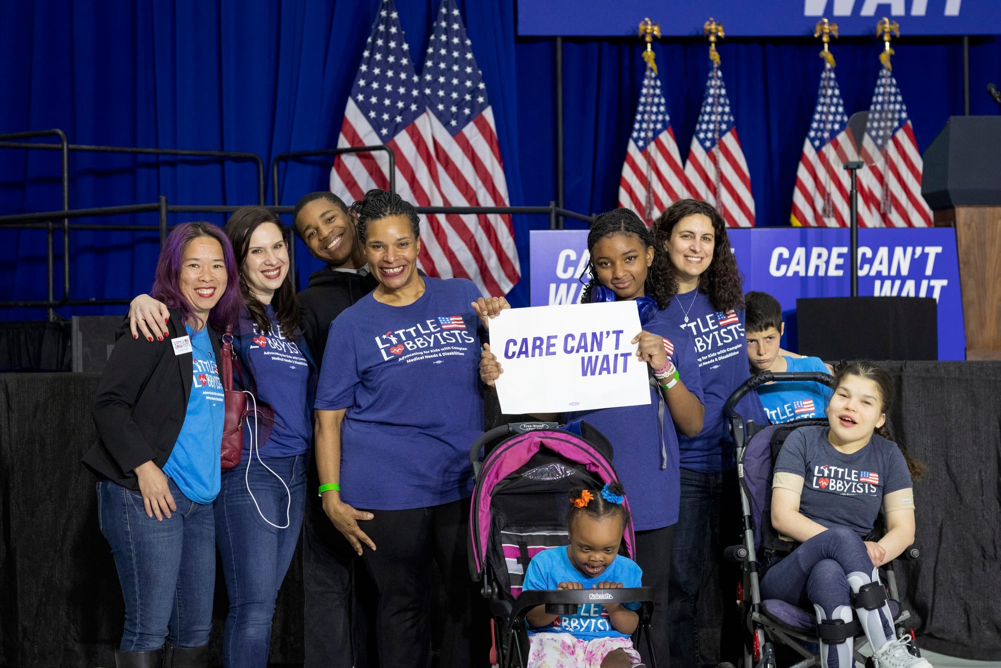 Another group of Little Lobbyists families poses in front of the stage, some standing, some using mobility devices. Danielle’s daughter Autumn holds a sign that says “Care Can’t Wait.”