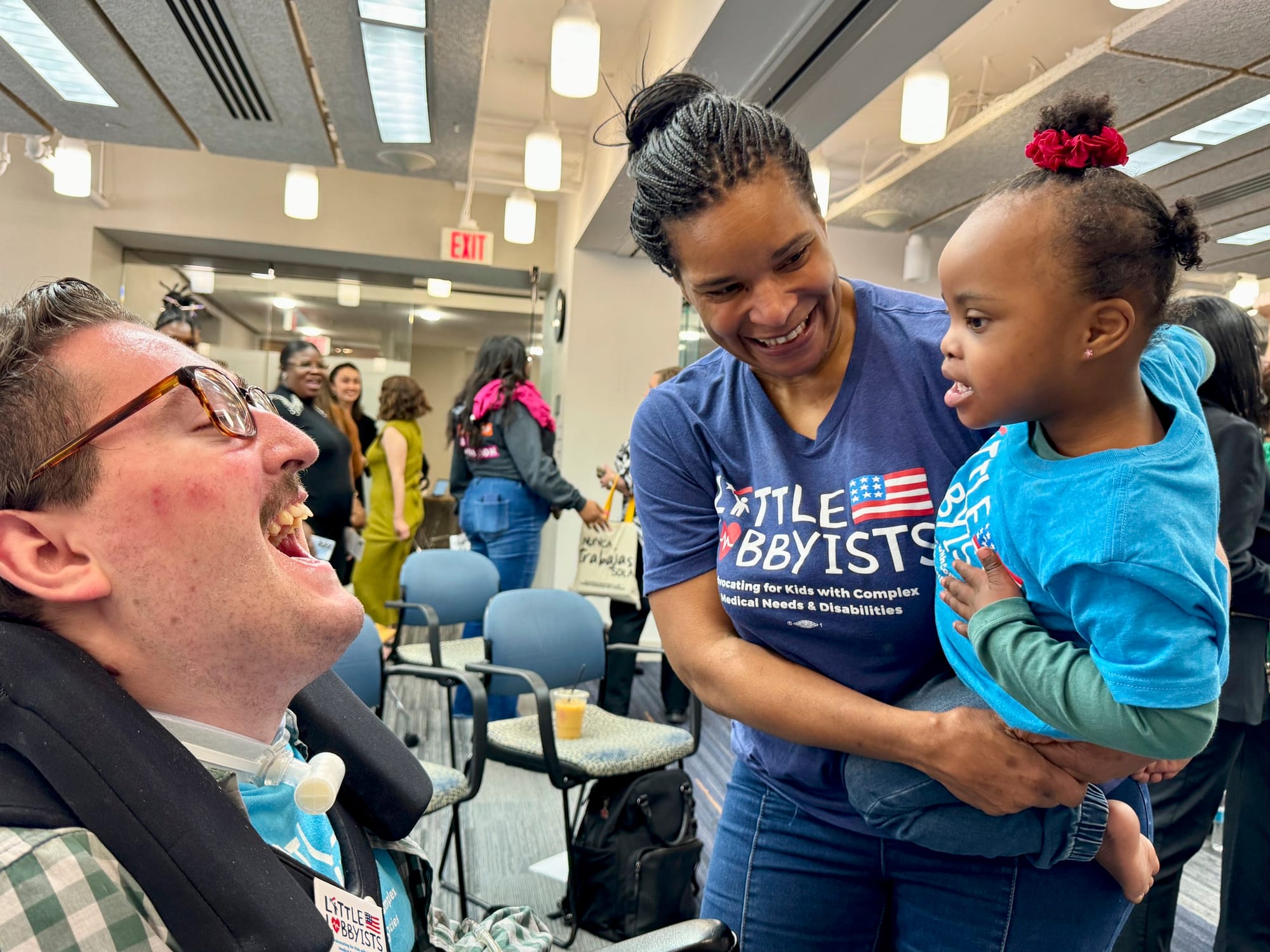 At the Congressional Town Hall, Rob smiles at Destiny, who is being held in Danielle’s arms. Destiny is giving Rob the once-over. They are in a large conference room with banks of rectangular overhead lights.