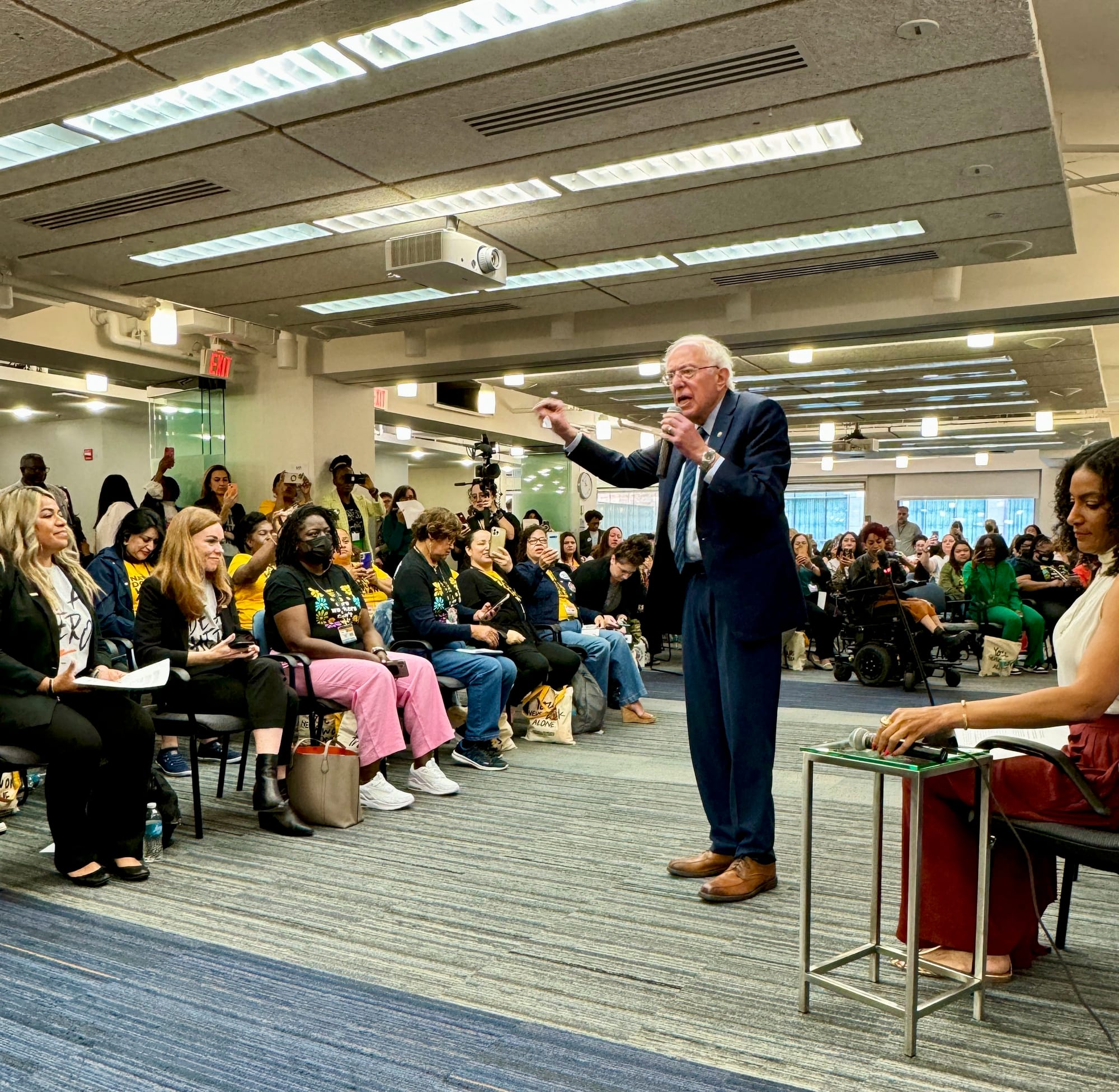 Senator Bernie Sanders, wearing a suit and with his trademark wild white hair, speaks energetically into a microphone at the Town Hall to a tightly packed audience who sit on folding chairs.