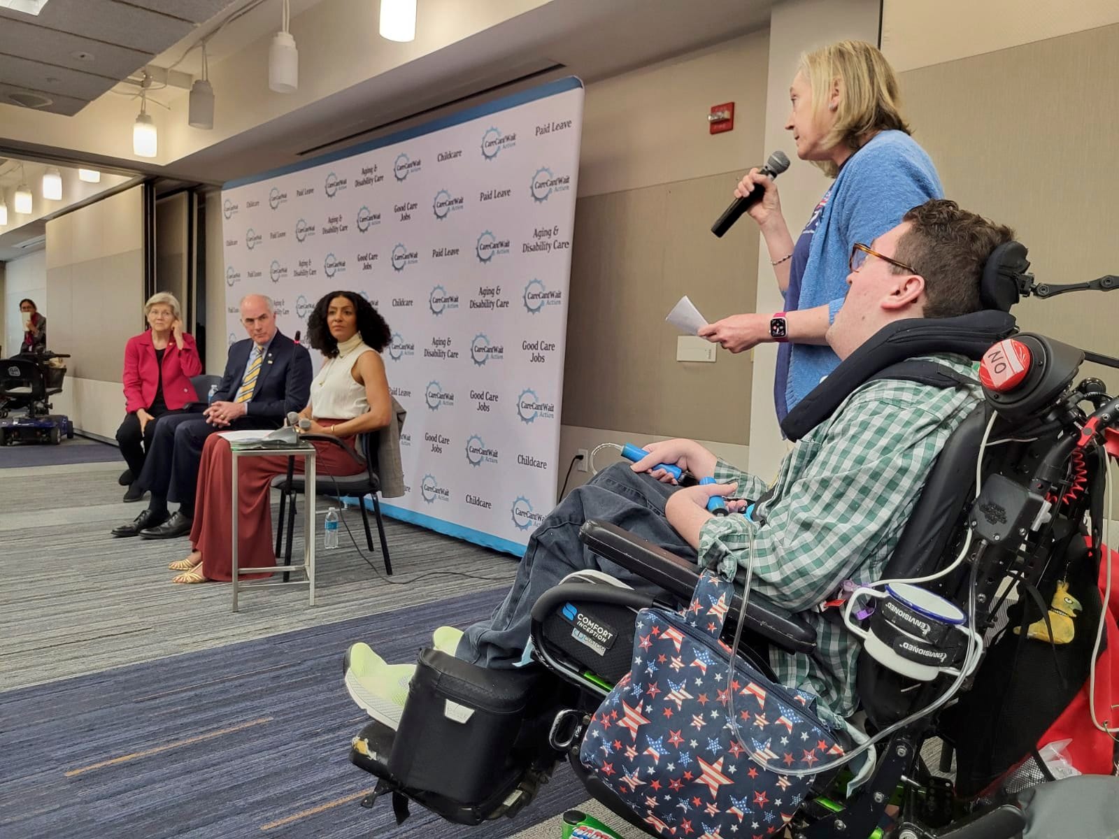 Jeneva holds a microphone, with Rob next to her in his wheelchair, in 3/4 view of their backs. She is asking a question of Senator Elizabeth Warren, who wears a red suit jacket and is seated next to Senator Bob Casey and the host Sarah Jones.
