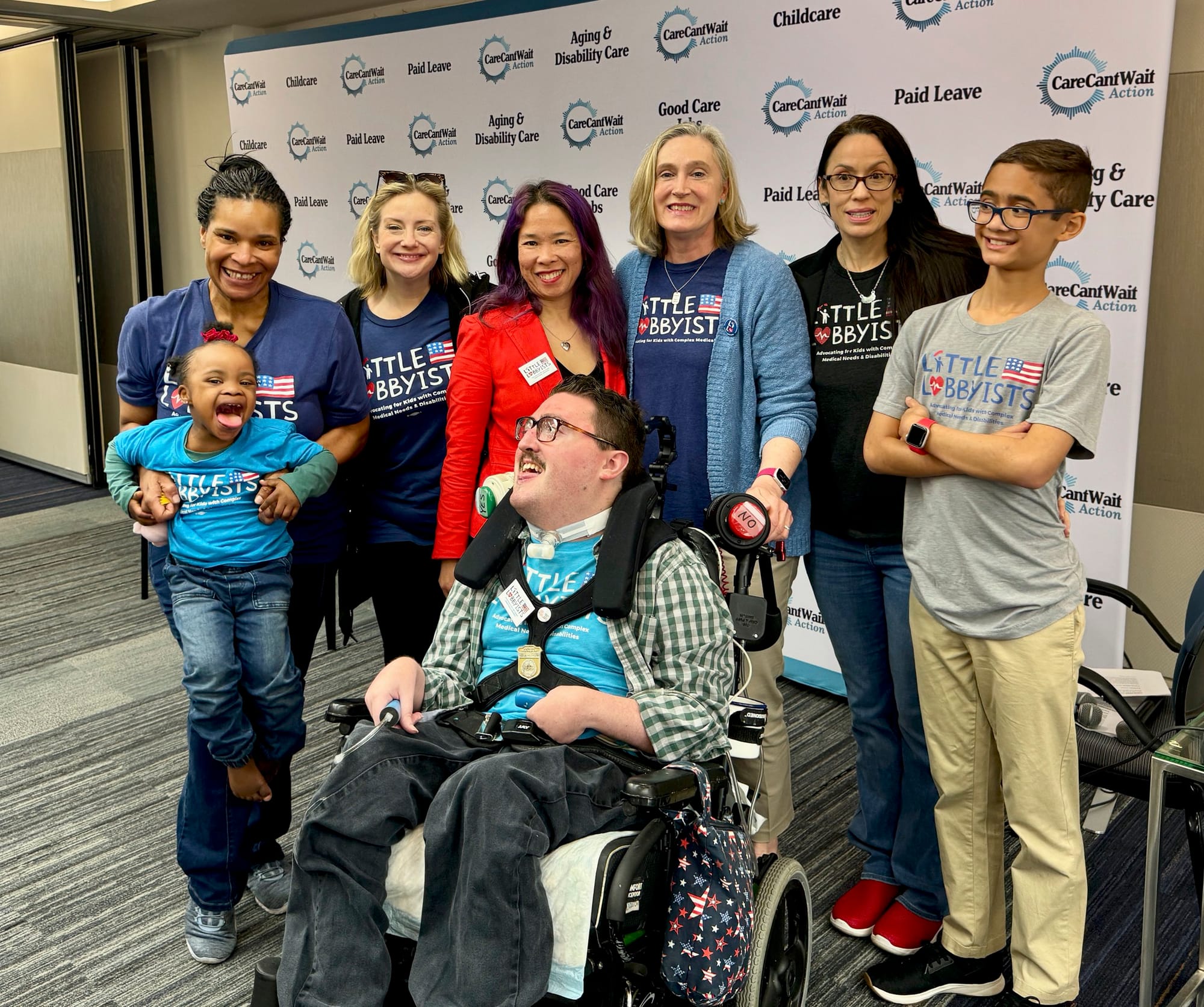 Our Little Lobbyists families pose in front of the Town Hall backdrop, white with the blue logo of the Care Coalition and various slogans about care. Rob is posed in his wheelchair at the front. From l to r: Danielle holding Destiny, Daya, Elena, Je