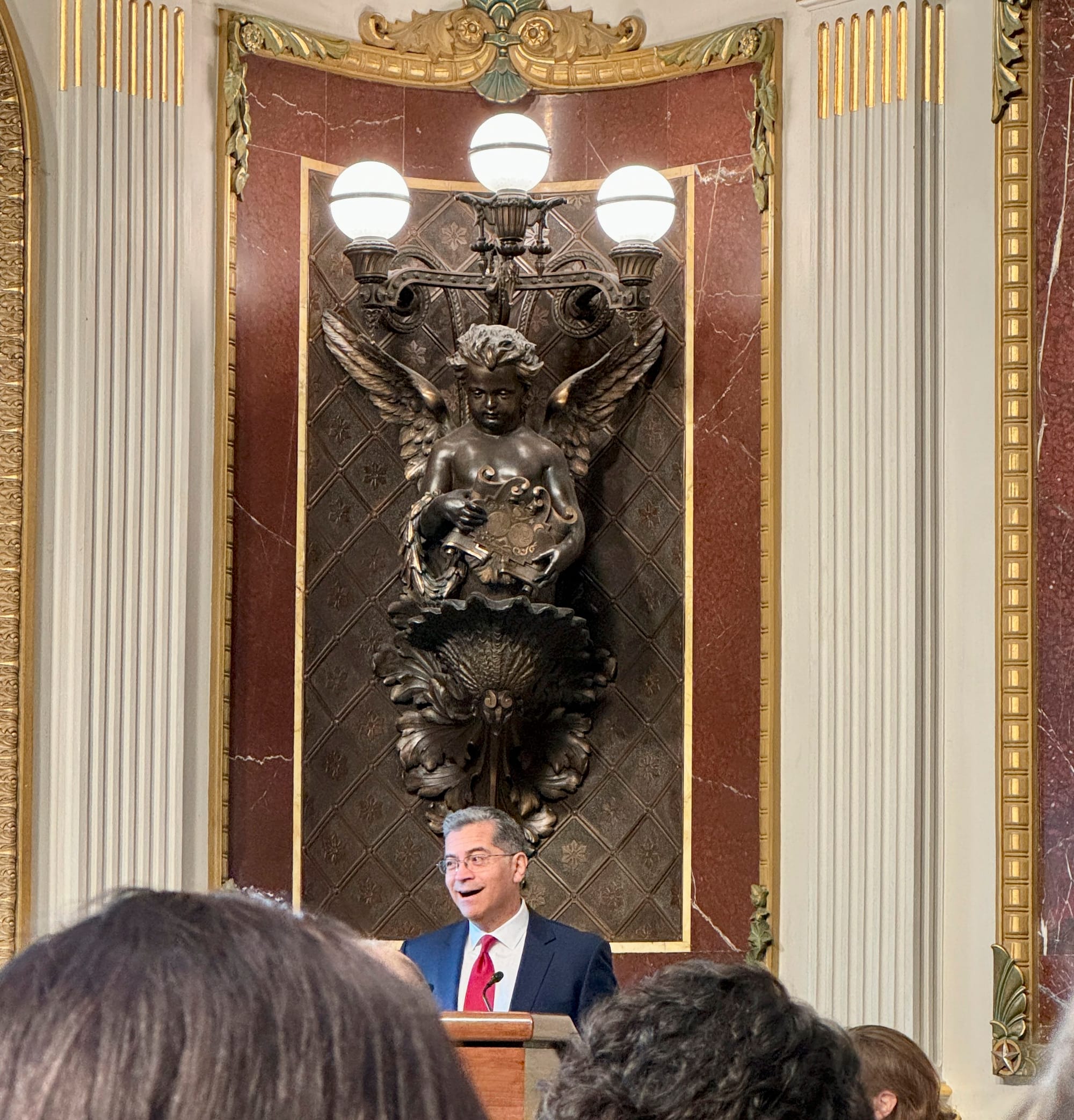 Secretary of Health and Human Services, Xavier Becerra speaks from a podium in an elaborately decorated 19th century meeting room in the Old Executive Office Building, which is adjacent to the White House. Behind him is a large bronze wall decoratio
