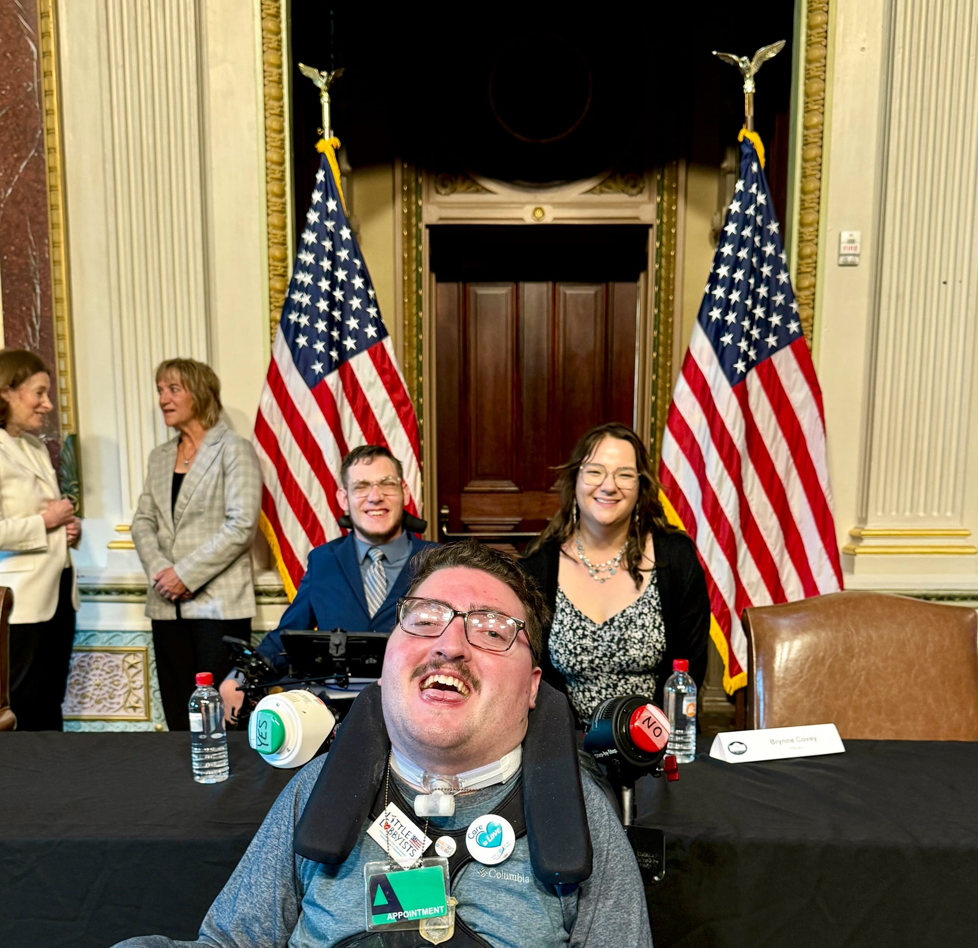 Rob poses in-between advocates Carson Covey (a fellow wheelchair- and AAC-user) and Carson’s sister Brynne. Behind them are two American flags flanking a decorative bronze doorway.