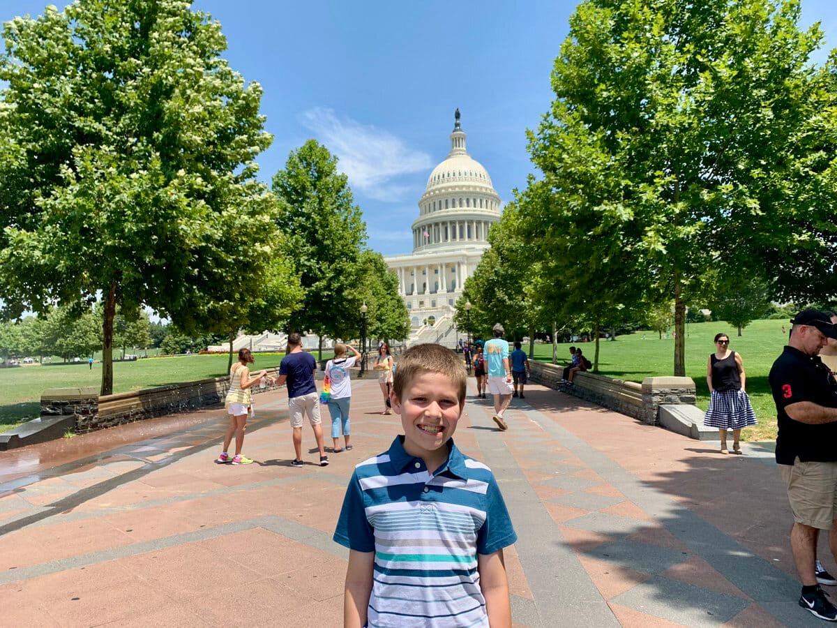 A young boy poses on a tree-lined street near the U.S. Capitol, with the Capitol dome in the background.
