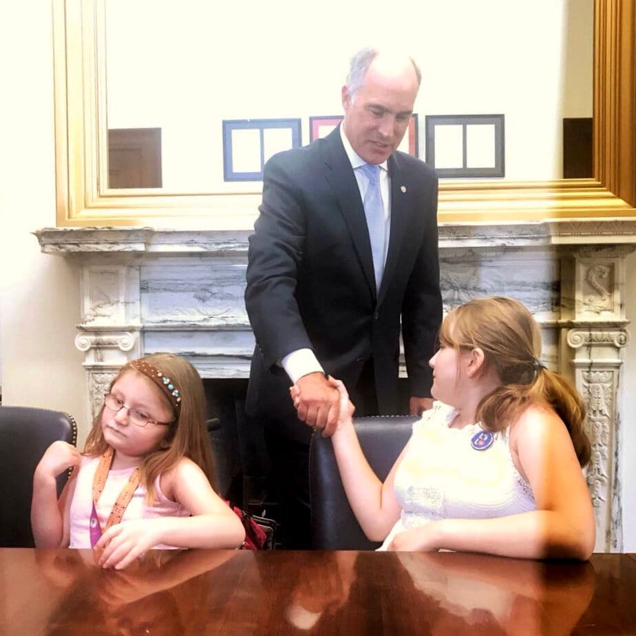 Two young girls are seated at a conference table in front of a marble fireplace. One shakes hands with her Senator.