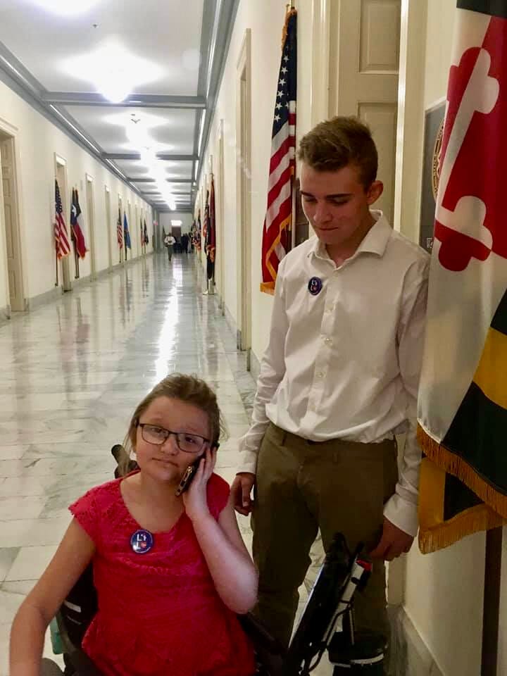 A young girl sits in her wheelchair and makes a phone call while her brother stands next to her. They are in a hall in a congressional office building.