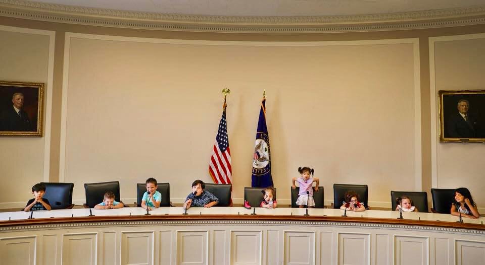 Several young children are seated at a long legislative dais, flanked by flags.