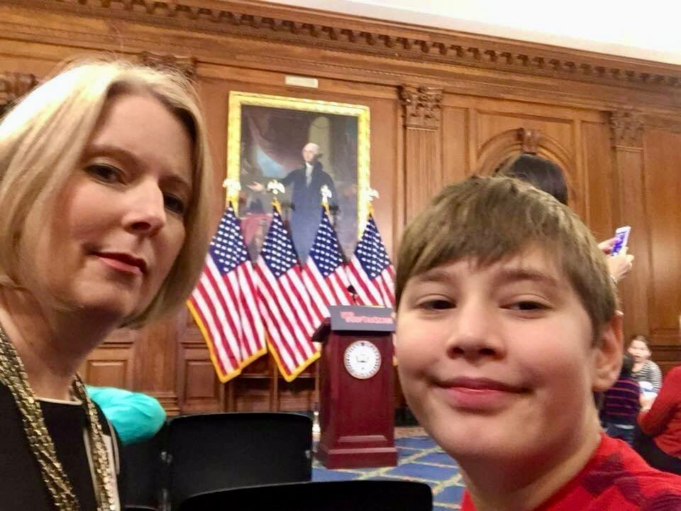 A mom and her son pose for a photo after a U.S. Senate press conference. The podium and U.S. flags are in the background.
