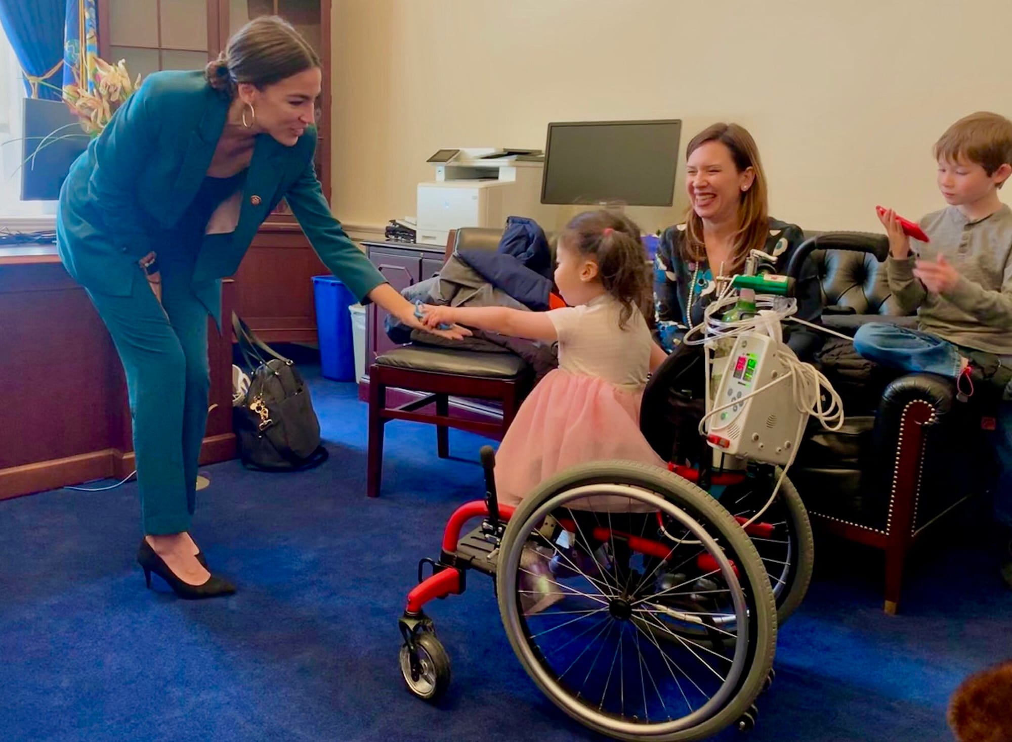 A new member of Congress wearing a vivid turquoise suit bends forward to grasp the hand of a Little Lobbyists child who reaches out to shake her hand.
