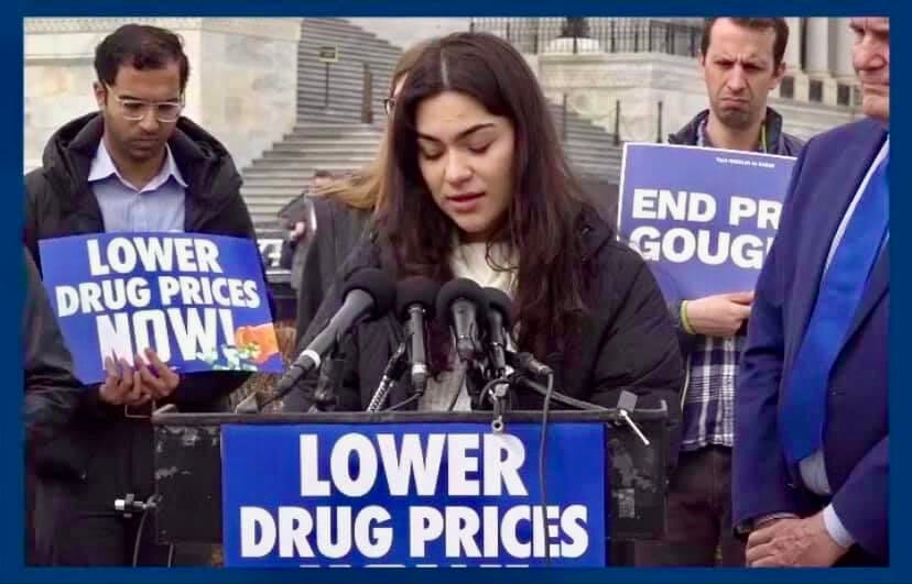 A young woman representing Little Lobbyists speaks at a podium marked with a “Lower Drug Prices” sign outside in front of the U.S. Capitol.