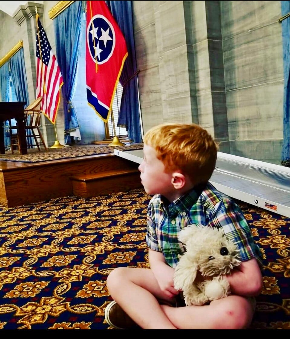 A young boy sits on a patterned carpet with state flags after a state legislative press conference.