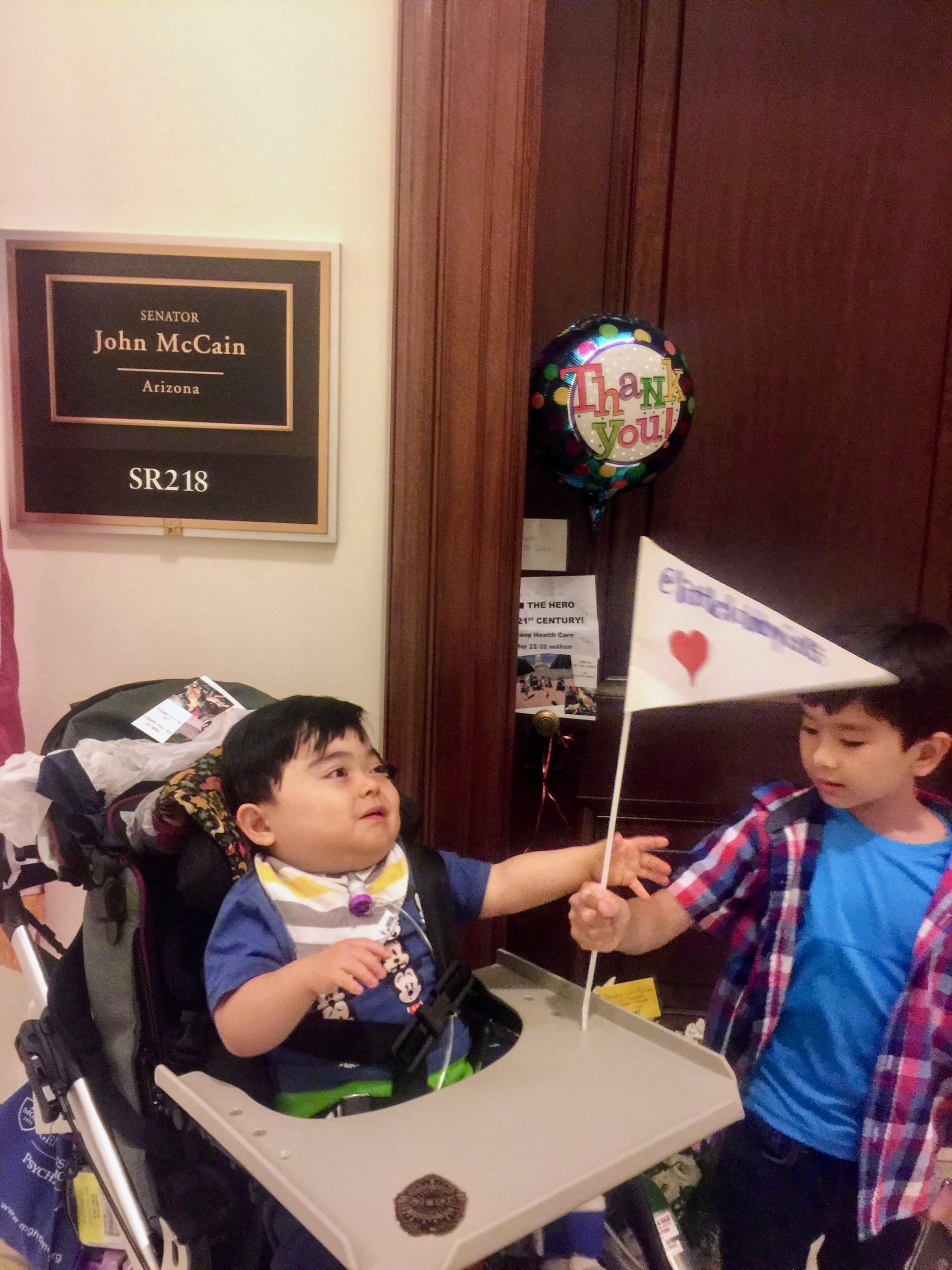 A young boy sitting in a wheelchair and his sibling stand in front of a congressional office door.