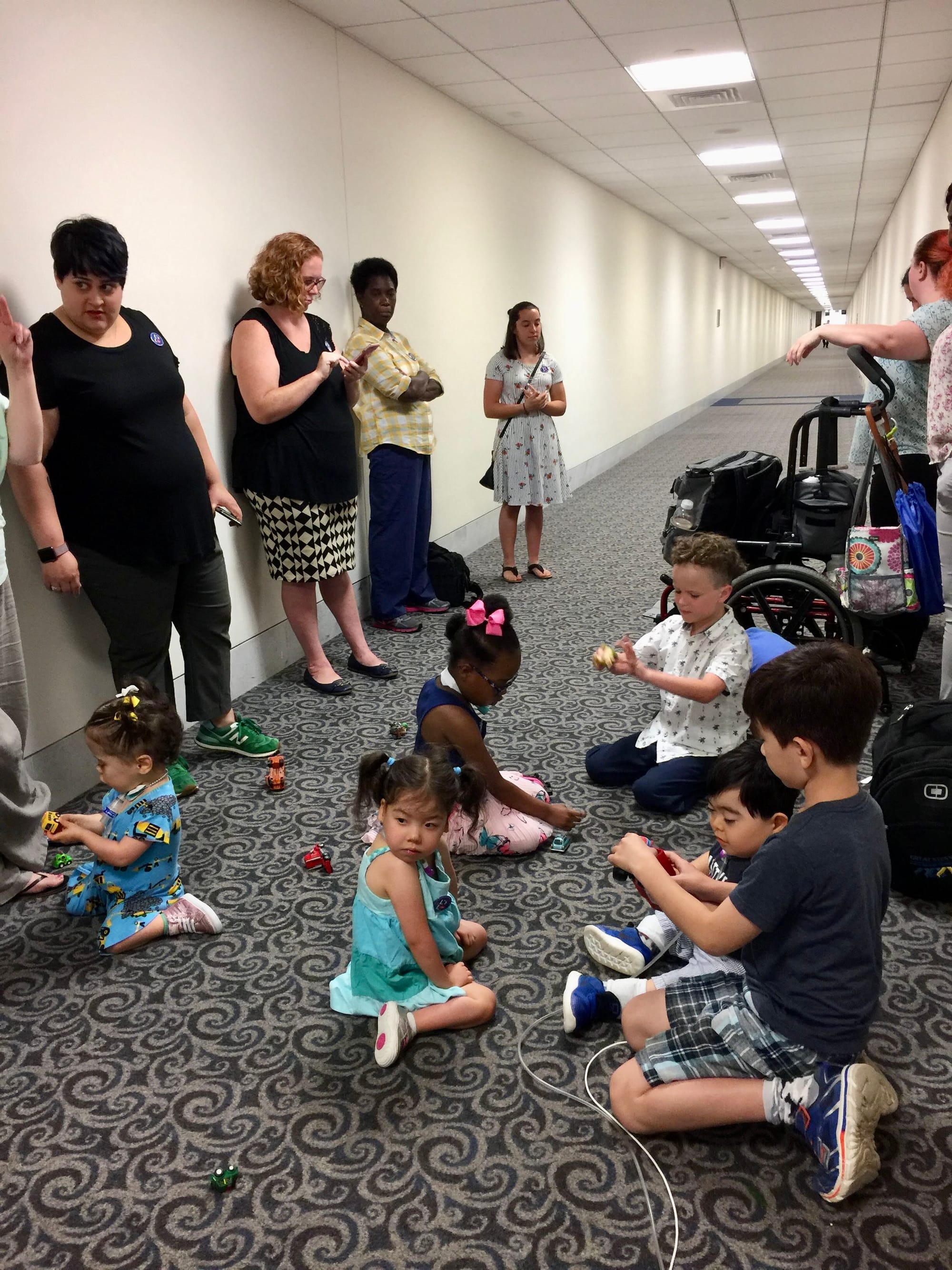 Little Lobbyists families sit and stand along a carpeted hallway in a congressional office building.