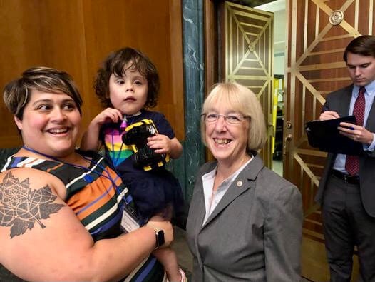 Standing before decorative doors in a congressional meeting room, a mom with a stunning tattoo on her arm holds her young daughter while posing with a U.S. Senator.