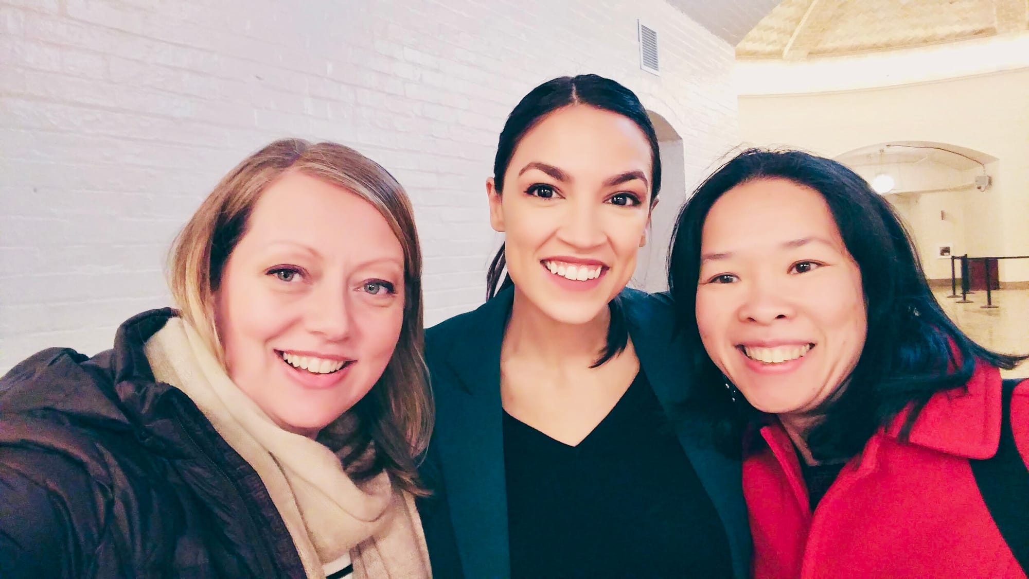 Two Little Lobbyists moms pose with a new member of Congress in a white-brick lined hallway.