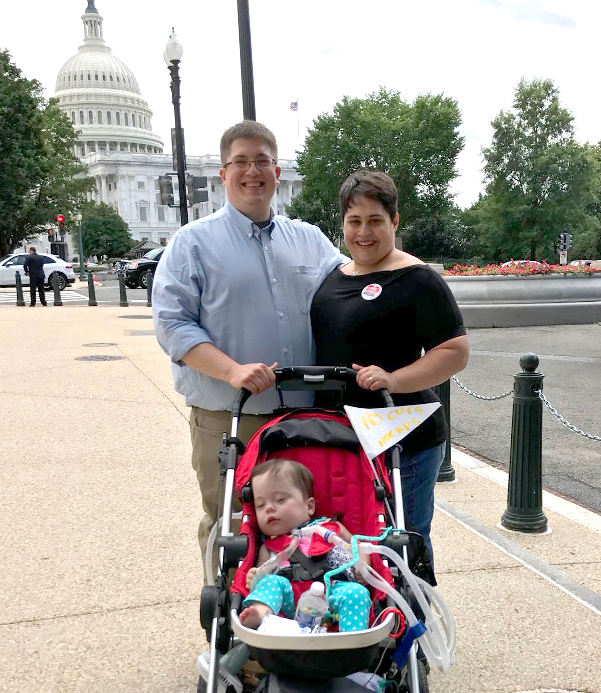 A family of three standing in front of the capitol building. The dad and mom stand proudly. The baby girl is in a red medical stroller and has a trach with ventilator tubes attached.