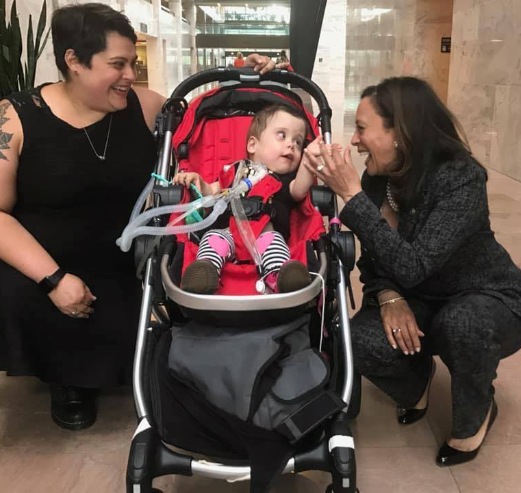 A woman in a black dress and vice president Kamala Harris crouch beside a baby in a red medical stroller. All three are smiling and the Vice President is holding the baby's hand.