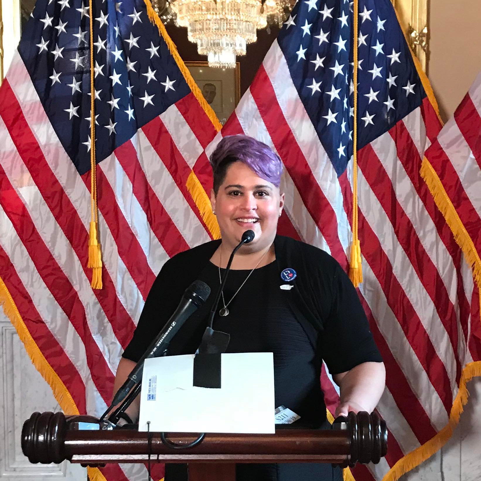 A woman in a black dress and purple hair with a smile standing in front of a podium in the Capitol.