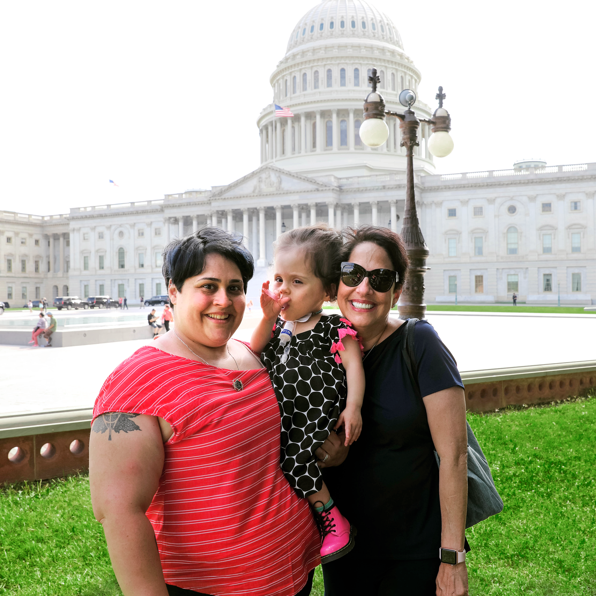 Two women hug a toddler in front of the Capitol building on a sunny day