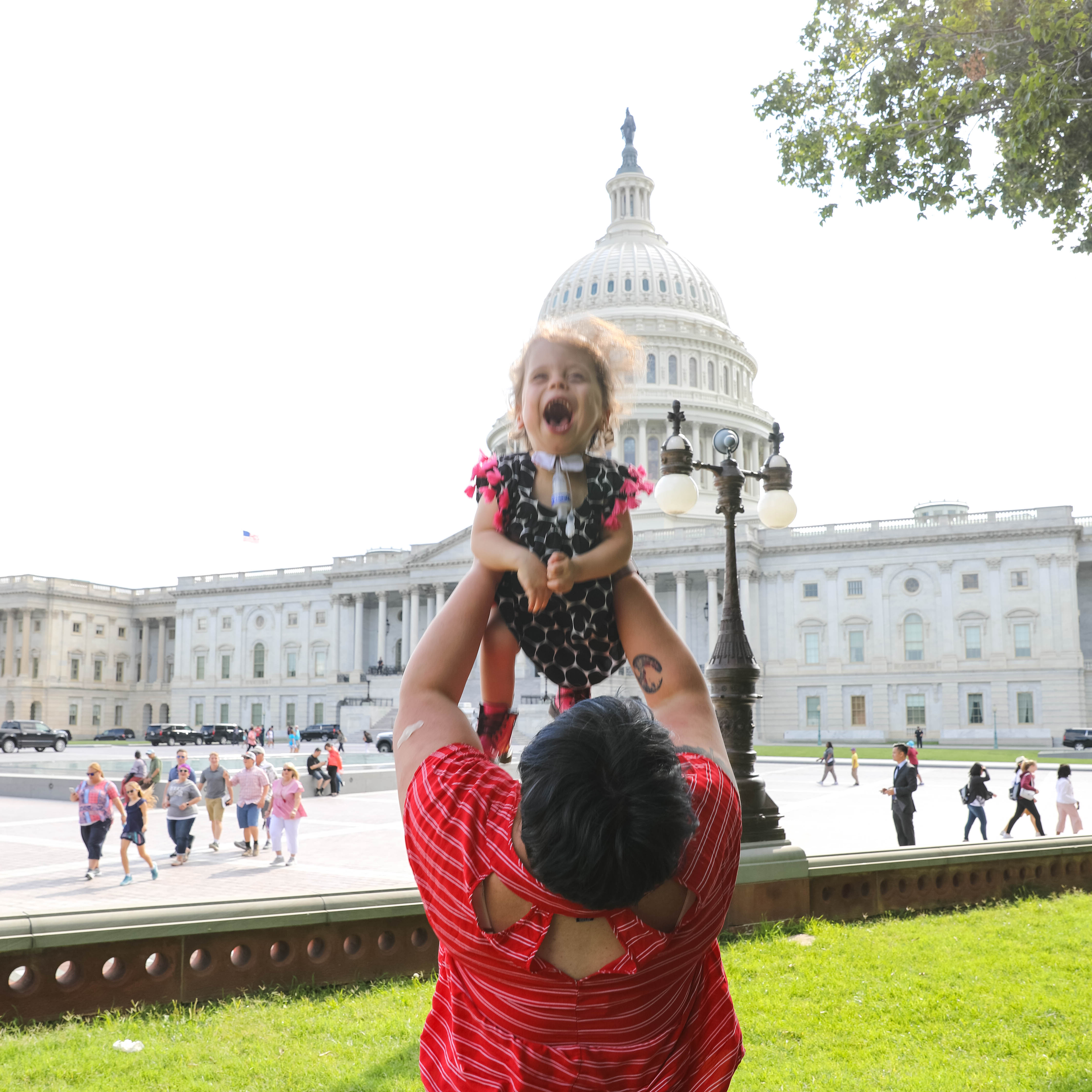 Baby girl with a trach wearing a pink and black and white dress has a gleeful expression as her mom lifts her high into the air in front of the Capitol building
