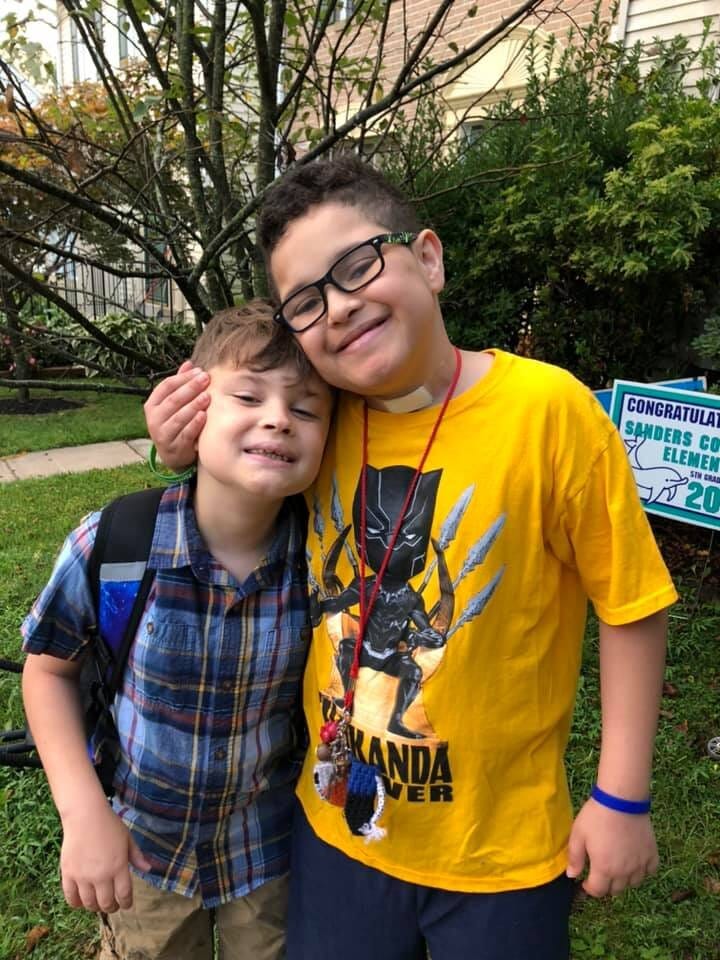 Kim’s sons Elijah (l) and Isaac (r). [image description: Two young boys with light skin stand closely together with their arms around each other. One wears a blue plaid shirt, the other a yellow t-shirt with the words “Wakanda Forever” and an image of Marvel Comics’ Black Panther.]