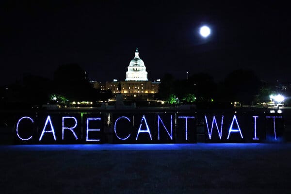 The U.S. Capitol Dome illuminated at night—in the foreground are a bank of pale blue lights spelling out “Care Can’t Wait.