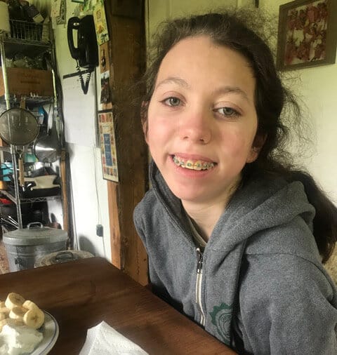 Elvina’s daughter Colby. [image description: A young woman with light-colored skin smiles at the camera; she is seated in a family kitchen. Her brown hair is pulled back from her face. She has braces on her teeth, and wears a gray hoodie.]